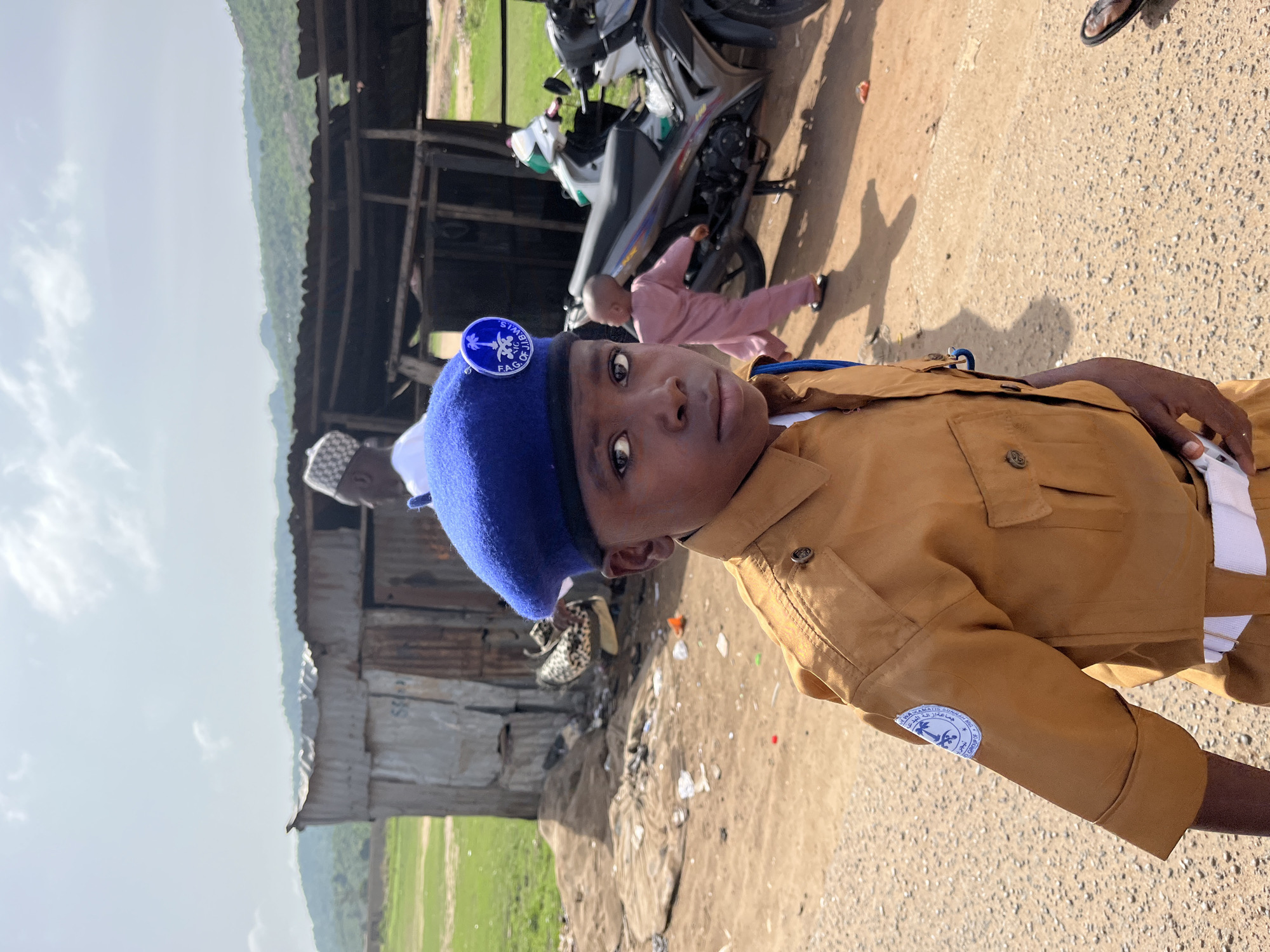 A boy in a tan uniform and blue beret with a round badge stands on a dirt road in a seemingly rural area. The badge on his beret has blue lettering around the edge, and the uniform sleeve has a round patch with Arabic lettering. In the background is a weathered, corrugated metal structure. A motorcycle is parked on the dirt surface, and another child in pink clothing is walking nearby. The ground is covered in debris. Beyond the structure is a green field, and a landscape with hills stretches into the distance under a light sky. The location is likely Jos, Nigeria based on the patch on the uniform.