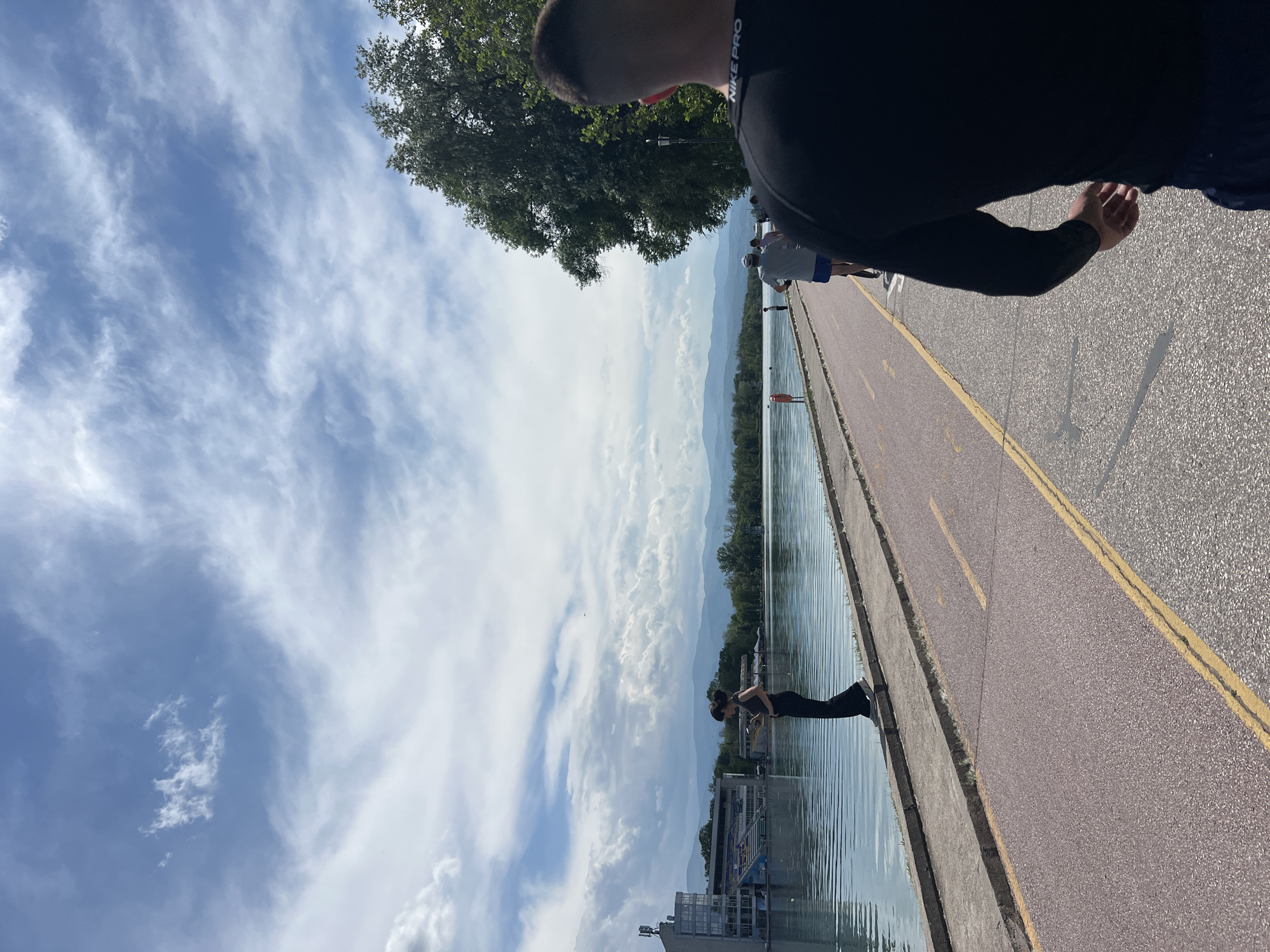 A medium-angle shot shows a horizontal scene alongside a body of water, likely the rowing canal in Plovdiv, Bulgaria. The image is taken from an elevated position on a paved walkway with a yellow line dividing it. A person in black attire stands at the water's edge, appearing to interact with the water. A tall building stands in the background to the left. Other people can be seen further along the pathway. The sky is filled with fluffy white clouds, and the horizon reveals distant mountains. A tree and the partial view of a person wearing athletic apparel with "NIKE PRO" are visible at the top right corner.