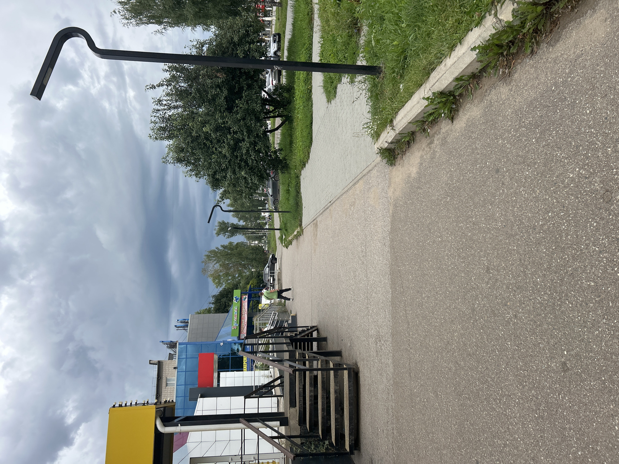 The image depicts a sidewalk and street scene under a cloudy sky, likely in Chaikovsky, Russia. In the foreground, a paved sidewalk runs parallel to a grey asphalt road. To the left of the sidewalk, a modern building features a yellow awning and a blue facade with red and white accents. A metal staircase leads up to the building's entrance. Further down the sidewalk, a person wearing a green shirt and dark pants walks away from the camera. Beyond the sidewalk, a green lawn with scattered trees lines the road. Several cars are visible parked or driving on the road in the background. A distinctive, modern black lamppost with a curved arm stands prominently on the right side of the image. The overall environment appears to be a commercial or mixed-use area within a city.
