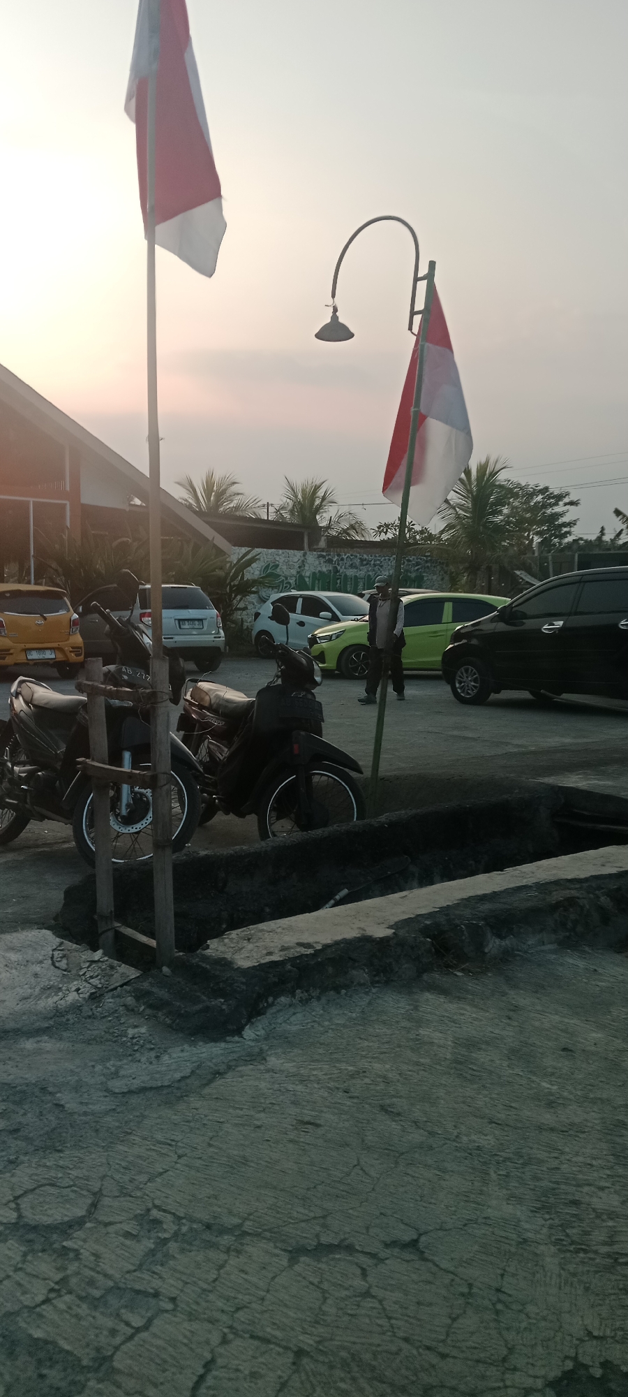 The image shows an outdoor scene in Banyuraden, Indonesia. In the foreground, two motorcycles are parked on a concrete surface. The motorcycles are black and have visible wheels and handlebars. Behind the motorcycles, there is a low, dark-colored wall or planter box.
In the middle ground, several cars are parked. There is a yellow car on the left, followed by a silver car, a white car, a lime green car, and a black car on the right. The cars appear to be in a parking lot.
Two individuals are visible in the middle ground. One person is standing near the lime green car, wearing dark clothing and a hat. Another person is partially visible further to the right.
The background includes a building with a slanted roof and palm trees. A street lamp with a curved arm is visible, and two flags, red and white, are mounted on poles. The flags are partially unfurled and appear to be Indonesian flags. The sky is a pale, hazy color, suggesting either early morning or late afternoon light. The overall environment appears to be a public area, possibly a parking lot in front of a business or establishment.