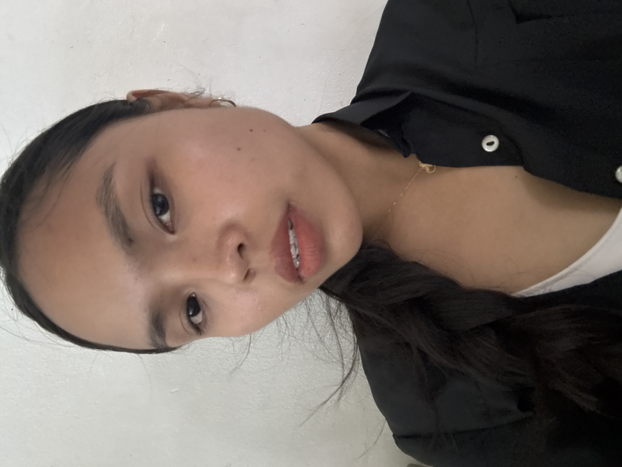 A young woman with dark hair, pulled back into a braid, is looking towards the camera. She is wearing a black collared shirt over a white top and a delicate gold necklace. The background is a plain, textured white wall.