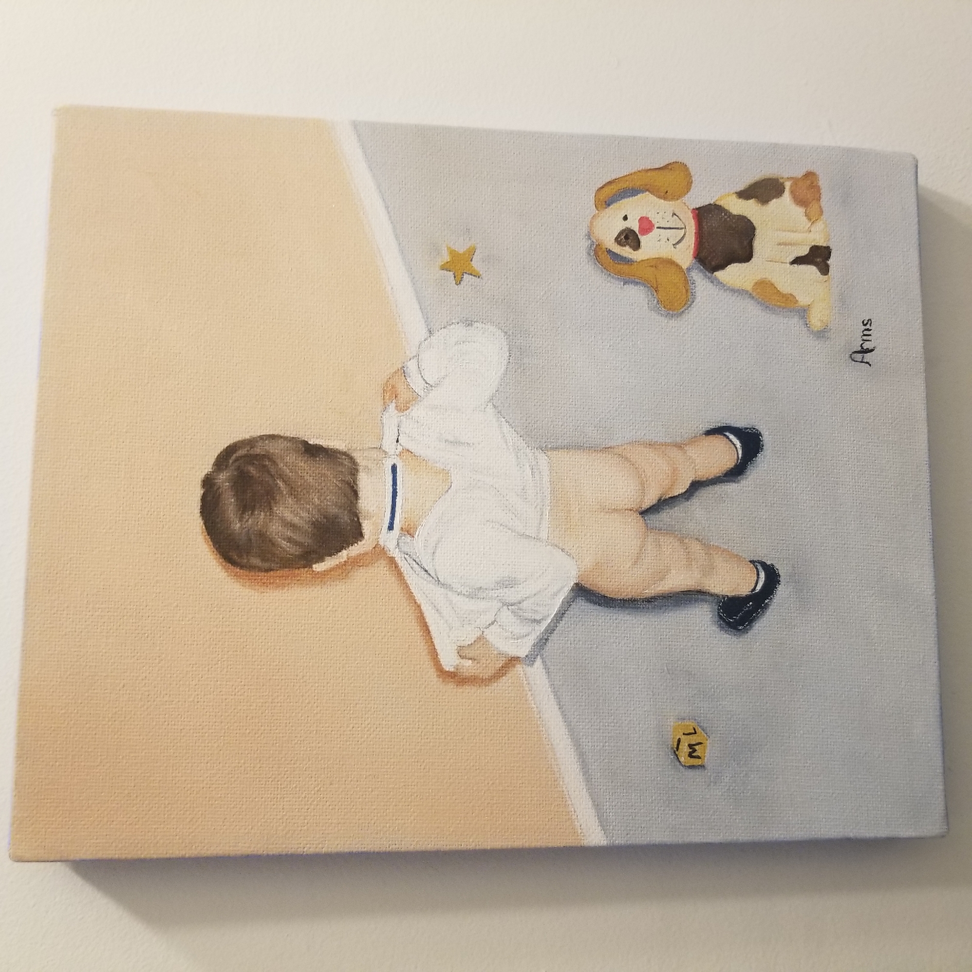 A canvas painting depicts a toddler with brown hair seen from the back, pulling a white shirt over their head. The toddler is wearing dark shoes with white trim. To the left of the toddler is a small yellow block with the letters "ML" in black. To the right of the toddler, a cartoonish tan and white dog with brown spots, a red collar, and a pink heart-shaped nose sits looking forward. A yellow star is painted on the wall behind the dog. The wall is a light peach color, and the floor is a light gray with a diagonal line suggesting a change in elevation or a corner. The painting is signed "Ams" in black cursive in the bottom right corner. The painting hangs on a white wall. This painting is located at the Children's Hospital of The King's Daughters in Norfolk, Virginia, which is part of the larger Hampton Roads region, near Virginia Beach, Virginia.