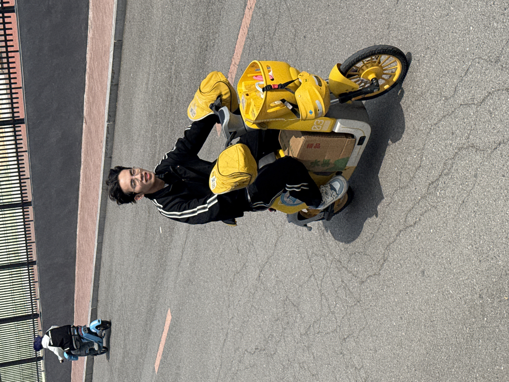 The image, oriented vertically, captures a bright outdoor scene with a young man on a yellow electric scooter. The man, positioned centrally, looks directly at the camera with a slight smile. He has dark hair, wears glasses, a black track jacket with white stripes on the sleeves, and black pants with similar stripes. His left foot is visible, wearing a light-colored athletic shoe.
He is seated on a bright yellow electric scooter, typical of shared mobility services. The scooter features a large yellow basket at the front, containing a red and white strap and some indistinct items. Two yellow hats, resembling delivery caps, are placed on the scooter: one on the handlebars and another on the rear section. Behind the man, a cardboard box rests on the scooter's back platform. Visible Chinese text on the scooter's body reads "美团" (Meituan), identifying it as belonging to a major Chinese delivery service. Another label above the box says "精品" (quality goods). On the cardboard box itself, the character "水" (water) is discernible.
The scooter is parked on an asphalt road with visible cracks, indicating wear. A faint orange painted line runs across the road surface. To the left, a dark-colored sidewalk or curb separates the road from a black metal fence or railing with horizontal slats. In the background, further down the road, another person is visible on a smaller, blue and black electric vehicle, resembling another shared scooter.
The scene is brightly lit by natural sunlight, casting distinct shadows under the scooters and the man, suggesting it is daytime with clear weather. The overall setting appears to be an urban or suburban street in Zhongtun, China.