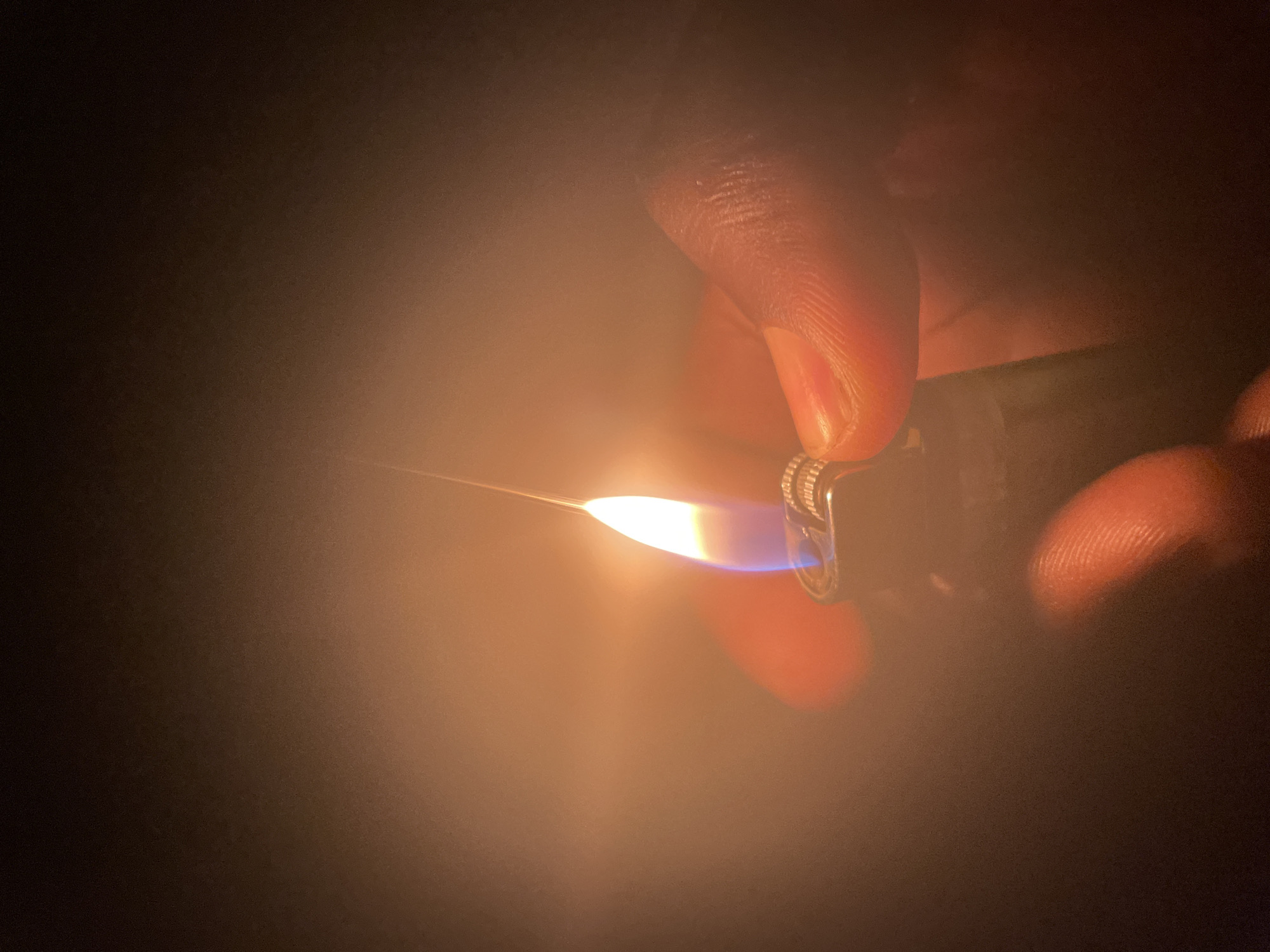 A close-up, low-light photograph depicts a dark-skinned hand holding a lit butane lighter. The flame is primarily white-yellow at the base transitioning to blue, and stretches horizontally to the left. The background is indistinct and dark. No specific details suggest a location in Jos, Nigeria.