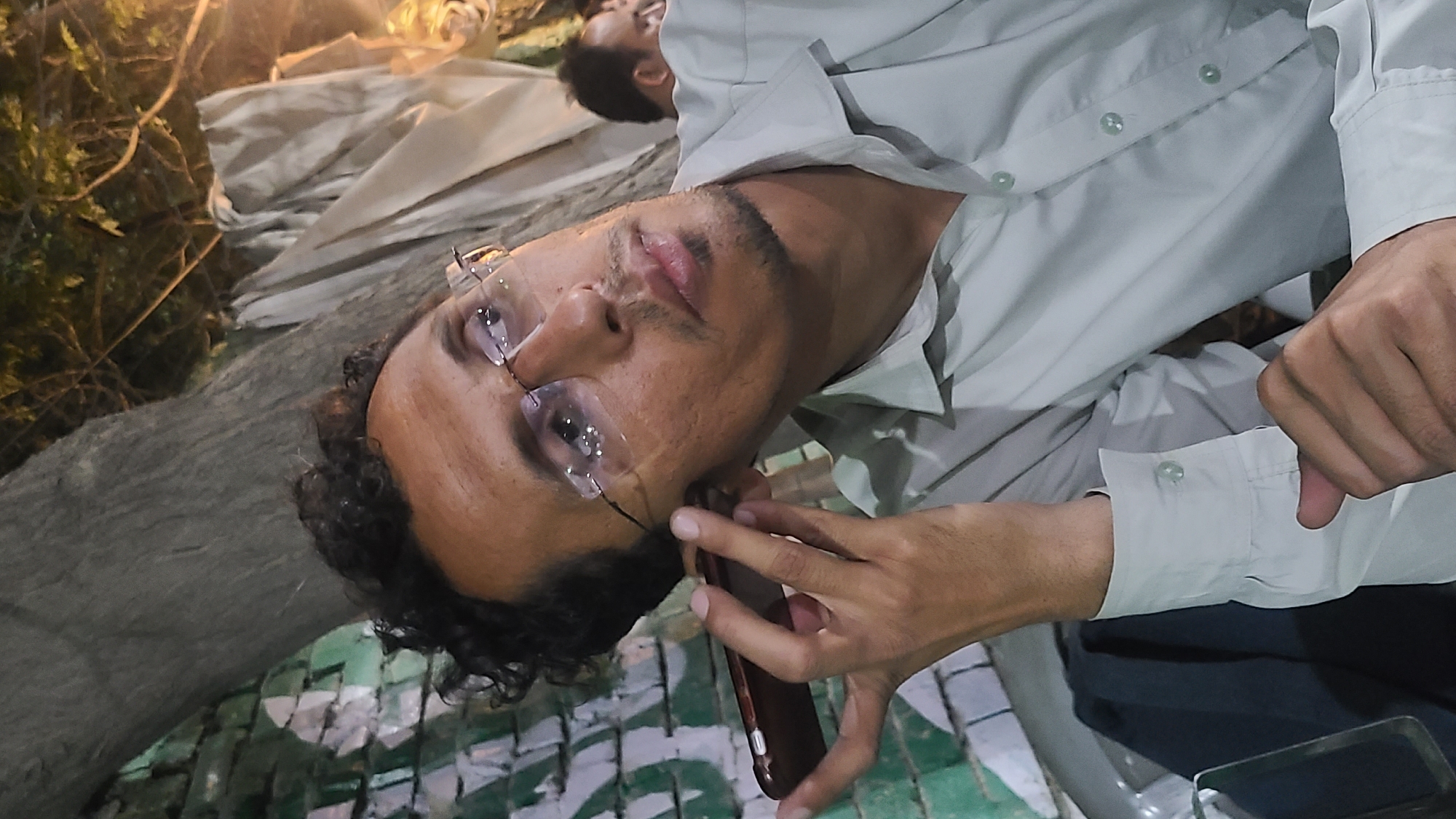 The image depicts a young man, likely in Quetta City Tehsil, Pakistan, at night or in the late evening. He has curly dark hair, a light mustache, and a short beard, and is wearing a light-colored, long-sleeved button-up shirt. He is wearing clear-lensed glasses with thin, dark frames that have reflective elements. The man is holding a dark-colored smartphone with a reddish-brown case to his ear, suggesting he is on a call. His expression is neutral as he looks forward.
In the blurred background, another individual, possibly a man, is partially visible, also wearing a light shirt. The setting appears to be outdoors, featuring green foliage and a tree trunk to the left. The ground beneath the main subject is adorned with a distinctive green and light-colored mosaic pattern. The scene is illuminated by artificial light sources, indicating the time of day. No text is visible in the image.