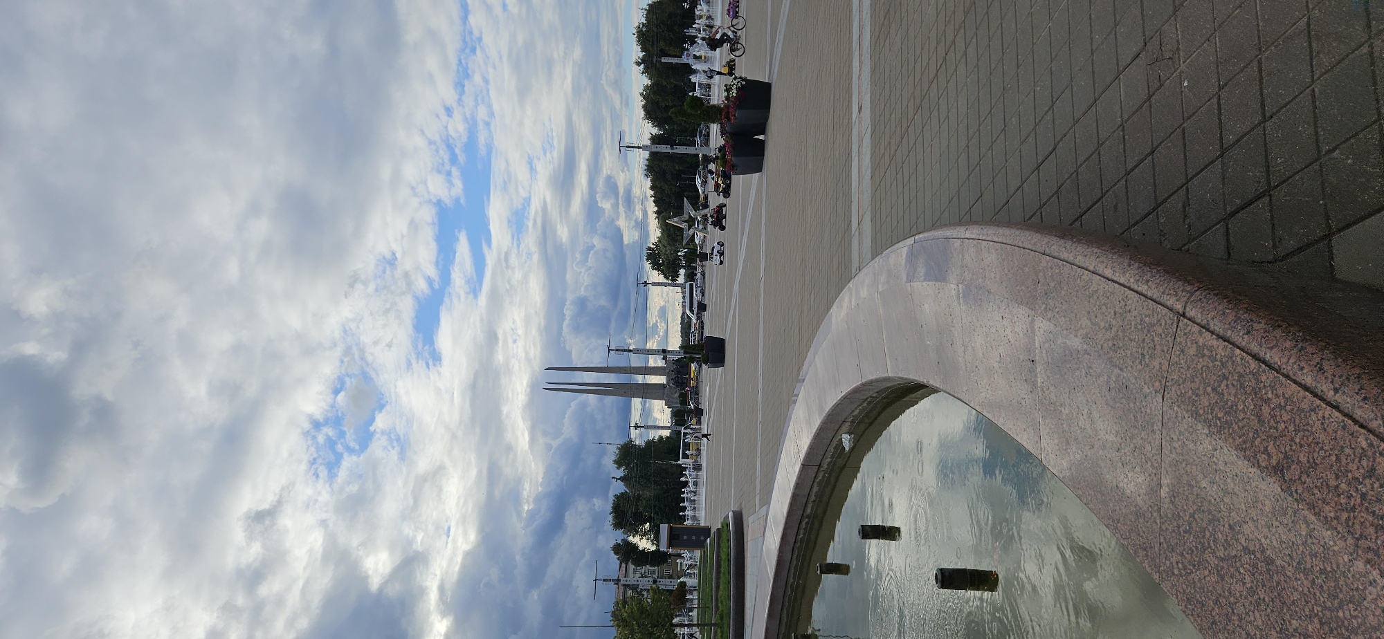The image depicts Victory Square in Vitebsk, Belarus. A large, curved fountain with dark cylindrical spouts is in the foreground, with its water reflecting the cloudy sky. The fountain's edge is made of polished reddish-brown granite. Beyond the fountain, a paved plaza stretches into the distance, laid with square grey pavers. A monument with three tall, tapering obelisks stands prominently in the middle ground. In front of the monument, there is a large, metallic star emblem on the ground. Several people are visible in the plaza, including a person walking near the fountain, individuals on various rideable toys such as small cars and a bicycle, and others in the distance. There are also several parked vehicles, including cars and vans. Trees line the perimeter of the square, and some buildings are visible in the background. The sky is filled with a dramatic mix of dark grey storm clouds and patches of bright blue sky.
