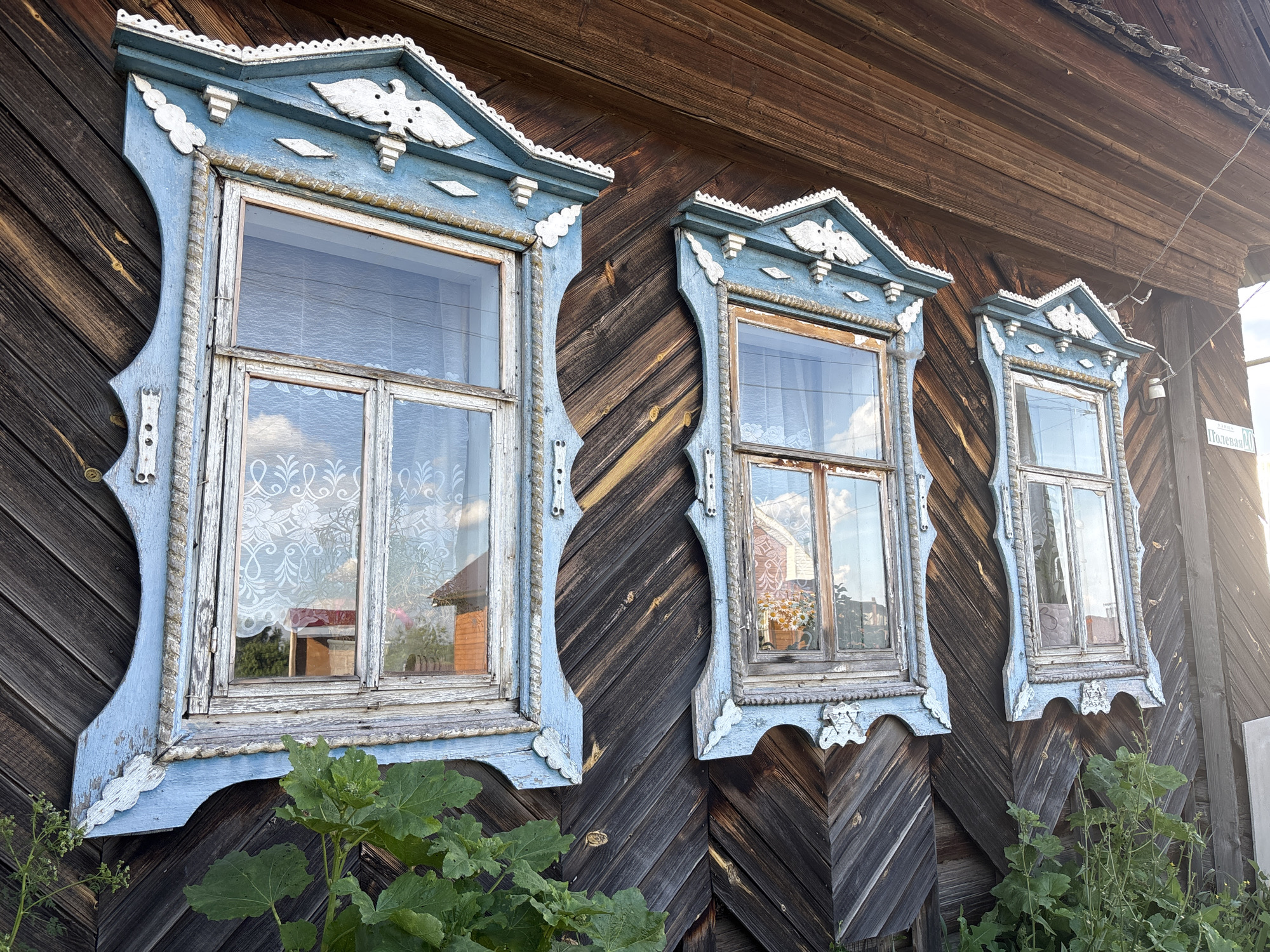 The image displays the exterior of a weathered wooden house in Niva, Russia. Three intricately decorated windows, framed by blue wooden trim with white detailing, are visible. The window frames are adorned with carvings of birds and geometric patterns, showcasing traditional Russian wooden architecture. The wooden siding of the house is dark and appears aged, with diagonal planks. Green foliage is growing around the base of the house, partially obscuring the lower portions of the windows. The windows reflect the sky and surrounding environment, suggesting it is daytime. A street sign with Cyrillic lettering is partially visible on the right side of the image. There are no people present in the scene.