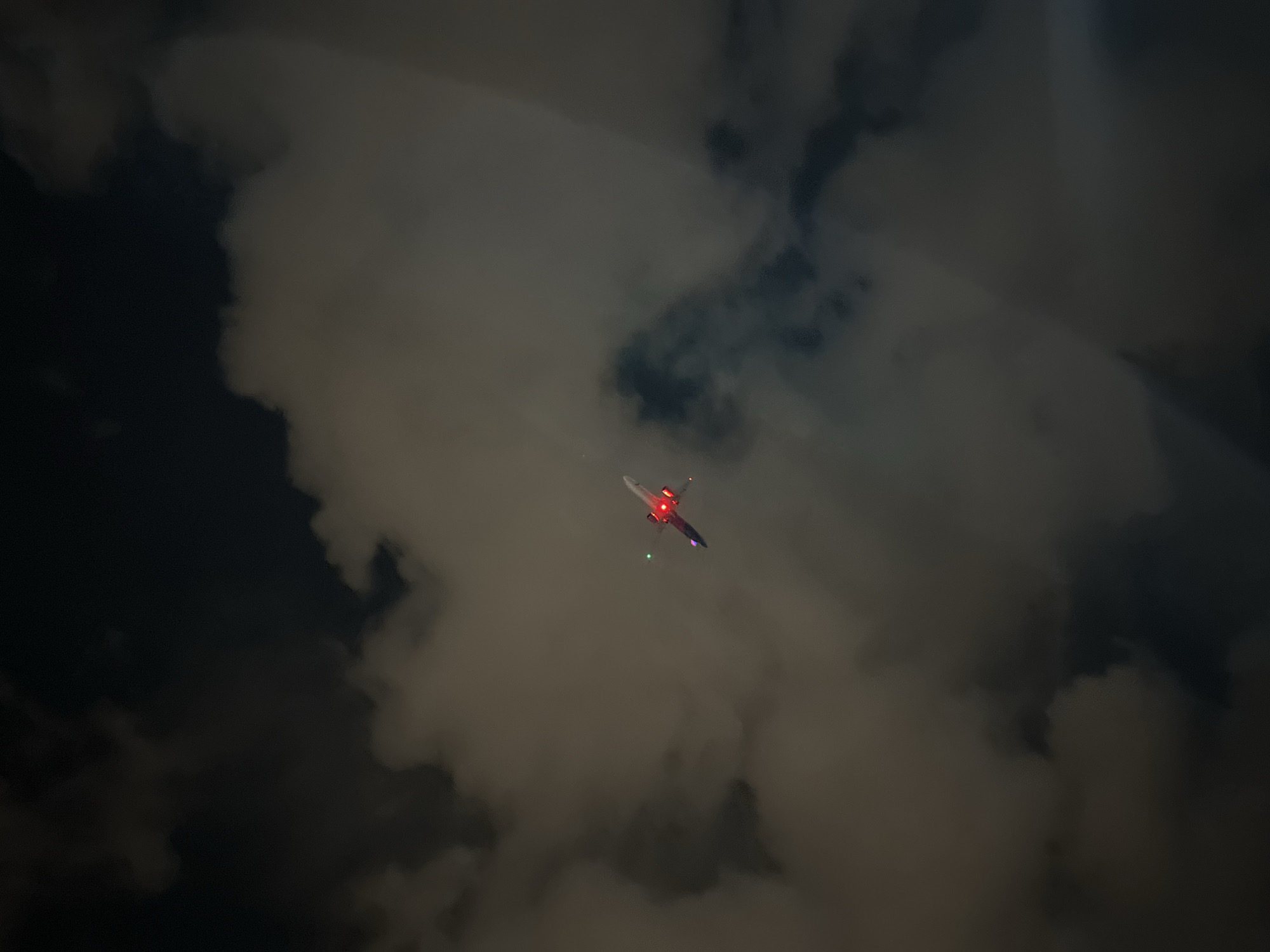 At night in Yerevan, Armenia, an airplane flies amidst dark clouds. The plane is angled away and its navigation lights are on.