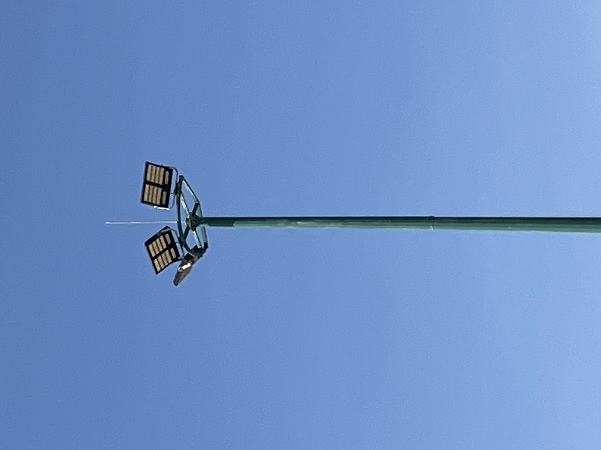 A tall, dark green metal utility pole stands vertically against a clear, bright blue sky in Bac Nha Trang Ward, Vietnam. At its apex, a multi-component lighting and power structure is affixed. This structure includes two large, rectangular units with dark frames and visible grid patterns, consistent with solar panels, mounted at an upward angle. Below these, and slightly to the left from the viewer's perspective, a single, conventional-style street lamp head extends horizontally and angles downwards. A thin, white or metallic rod, likely an antenna or lightning rod, extends vertically from the very top center of the entire assembly. The pole and its fixtures are well-defined against the unblemished sky, indicating bright daylight conditions. No people, vehicles, or other ground-level objects are visible within the frame.