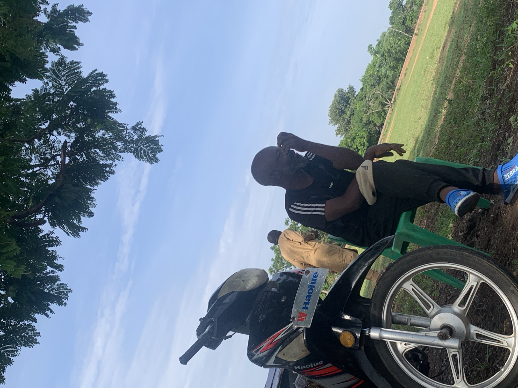 The scene appears to be in an outdoor area with a grassy field and trees, possibly on the outskirts of Akwanga, Nigeria. In the foreground, there is a black and red "Haojue" brand motorcycle with a license plate bearing the same name. The motorcycle is angled towards the camera. A man is sitting on a green plastic chair behind the motorcycle. He is wearing a black Adidas t-shirt with white stripes, black pants, and blue running shoes. He is touching his face and appears to be in thought. In the background, another person wearing a brown garment is standing near a grassy area. The sky is visible with some clouds.