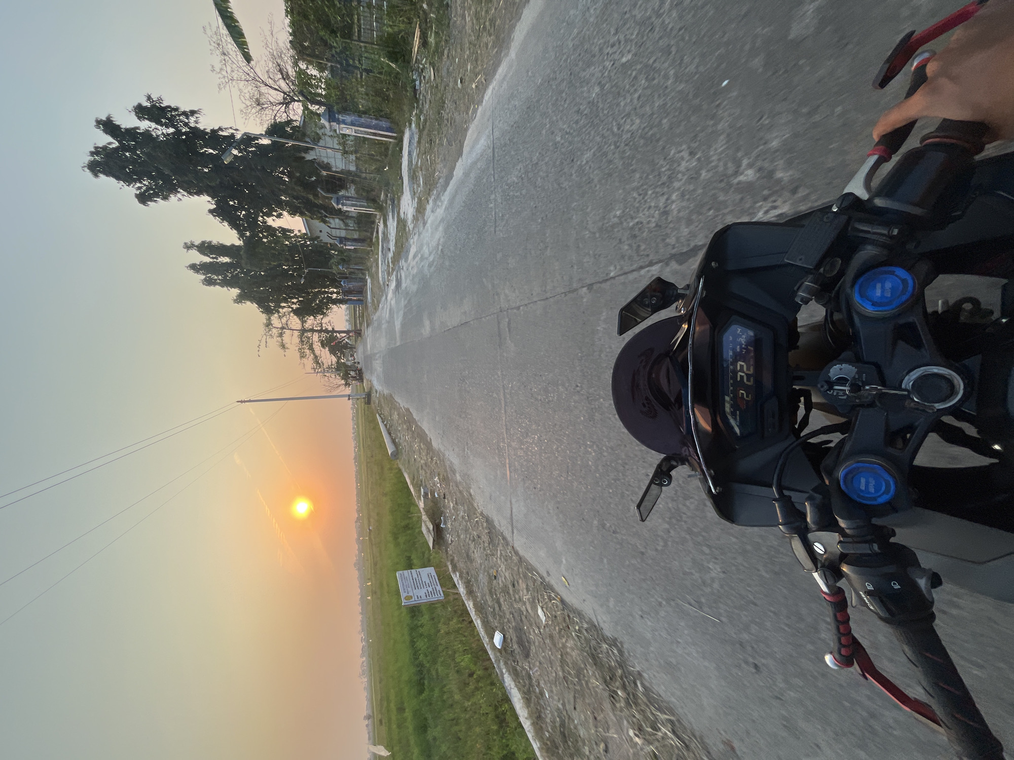 The image captures a first-person perspective of riding a motorcycle on a paved road in Pecangaan, Indonesia, during sunset. The sun is low on the horizon, casting a warm orange glow across the sky and illuminating a vast green field on the left side of the road. Tall, slender trees line the right side of the road, behind a fence and some buildings. The road itself is gray and appears to be in moderate condition with some cracks. In the foreground, the handlebars, digital dashboard, and a hand gripping the right handlebar of the motorcycle are visible. The dashboard displays numbers, indicating the speed or other metrics of the motorcycle. The overall scene suggests a peaceful journey through a rural or semi-rural area at dusk.