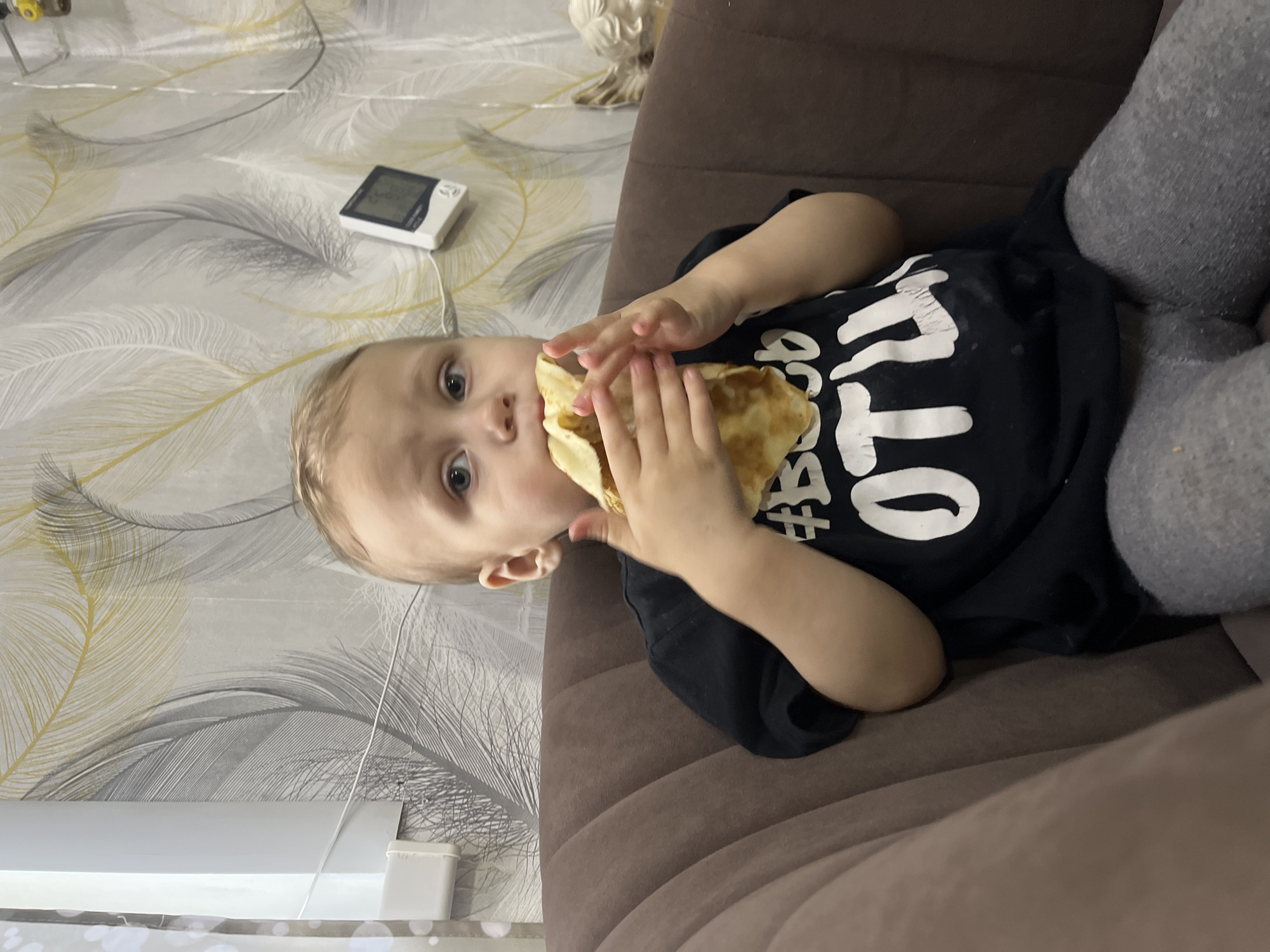A toddler is lying on a brown sofa, eating a folded pancake. The child is wearing a black T-shirt with white lettering and gray pants. The sofa is positioned against a wall with feather-patterned wallpaper. A white electronic device is on the wall near the child's head. The scene is indoors. The location is Фоки, Russia.