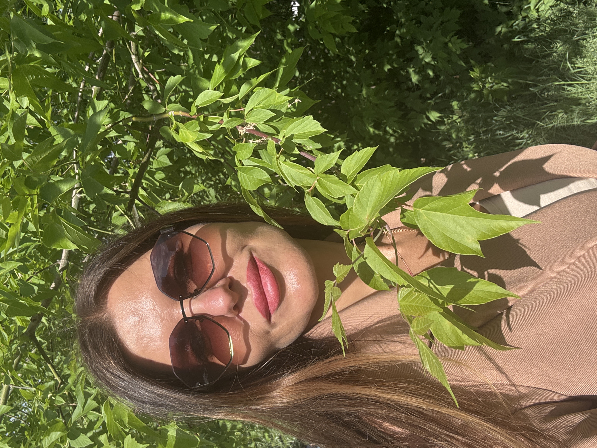 A medium close-up shot captures a woman from the chest up, positioned slightly to the left of the frame, with her head tilted. She is outdoors, surrounded by lush green foliage that forms the background and partially obscures her. The woman has long, brown hair that falls over her shoulders. She is wearing large, dark, geometric sunglasses that reflect the surrounding greenery and sky. Her lips are painted a vibrant pink, and she appears to be smiling slightly. She is dressed in a light brown blazer, revealing a glimpse of a cream-colored top underneath. A delicate chain necklace is visible at her collarbone. The sunlight casts dappled shadows across her face and clothing, indicating a bright, sunny day. The scene suggests a casual, outdoor setting, possibly a park or garden, within the city of Chaikovsky, Russia.