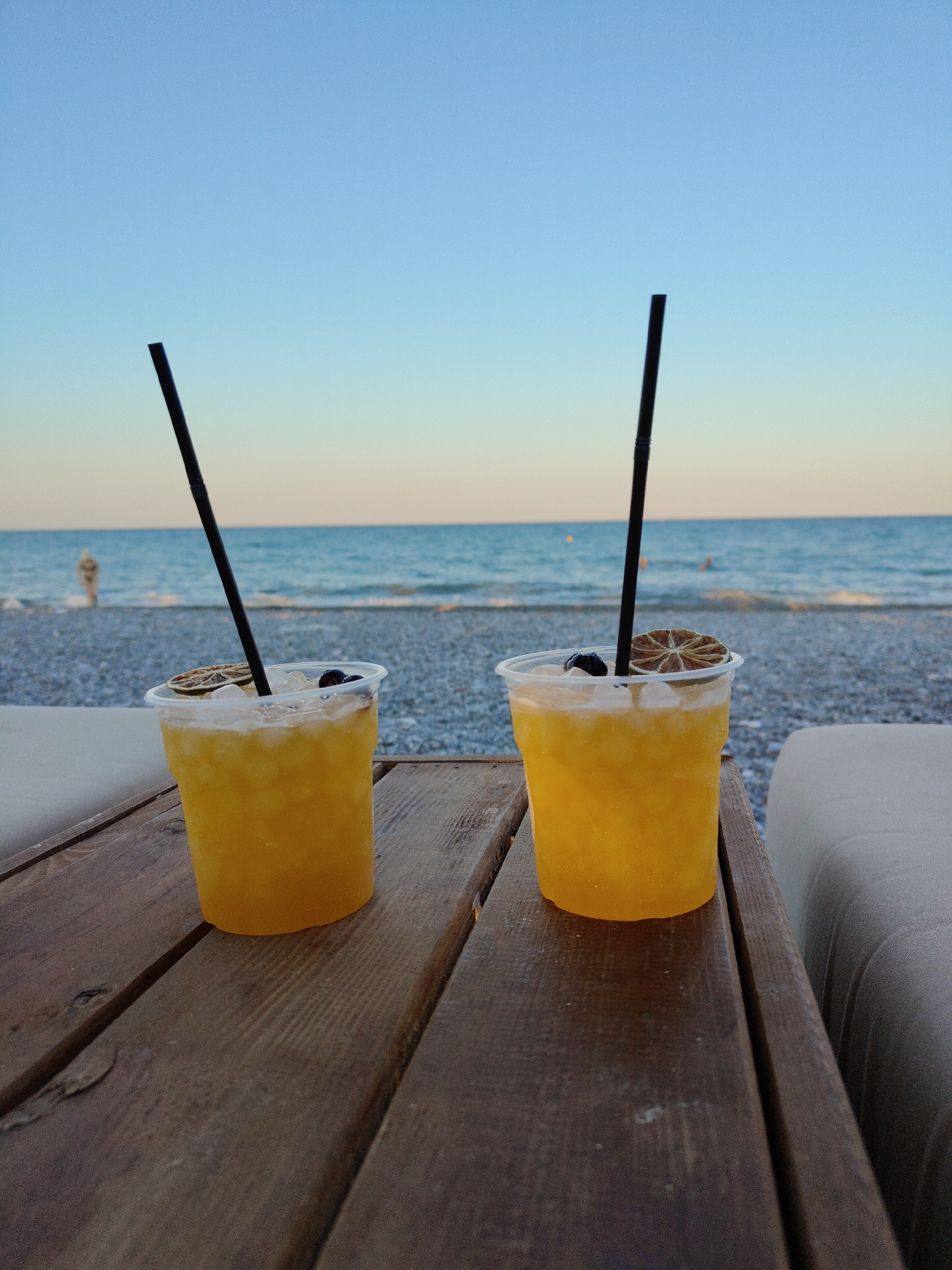 Two clear plastic cups containing bright yellow beverages with ice, black straws, a slice of dried citrus, and dark berries are placed on a wooden table. The table is set against a backdrop of a pebble beach and the ocean under a clear blue sky. The overall scene suggests a relaxing outdoor setting, likely a beachfront bar or cafe in Oraiokastro, Greece, given the specific location detail provided.