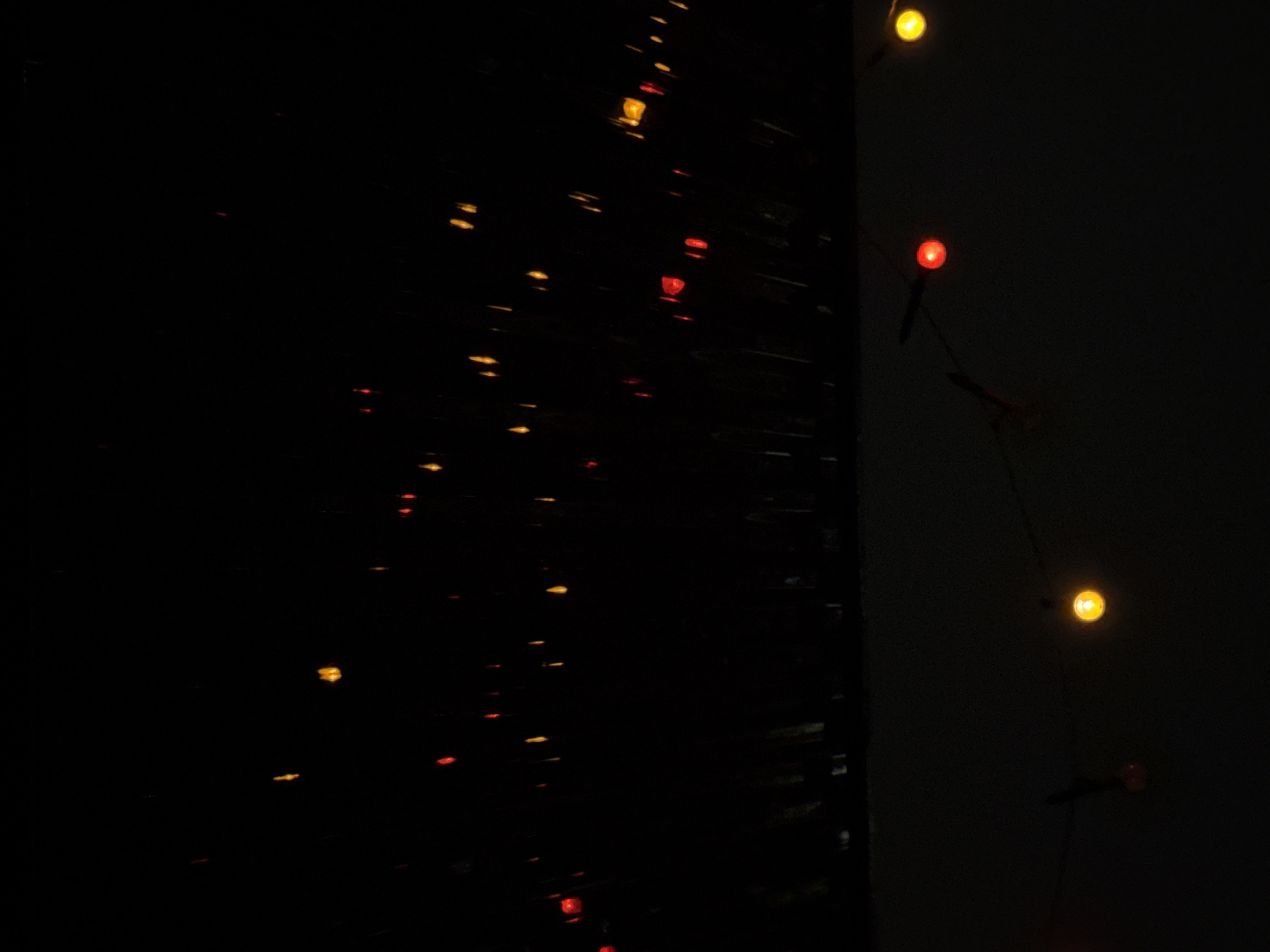 The image displays a dark scene with scattered red and yellow lights, suggesting they are Christmas lights or similar decorative illumination. The lights appear to be strung across a wire, with some lights hanging lower than others. The background is predominantly black, with some indistinct vertical shapes visible in the upper portion of the image, possibly representing a roofline or some form of overhead structure. The lower part of the image shows a lighter, muted surface, which could be a wall or building exterior. The overall impression is of a dimly lit outdoor setting at night. There are no people, animals, or specific identifiable landmarks present in the image, and no context to place it in Vostok, Kyrgyzstan.