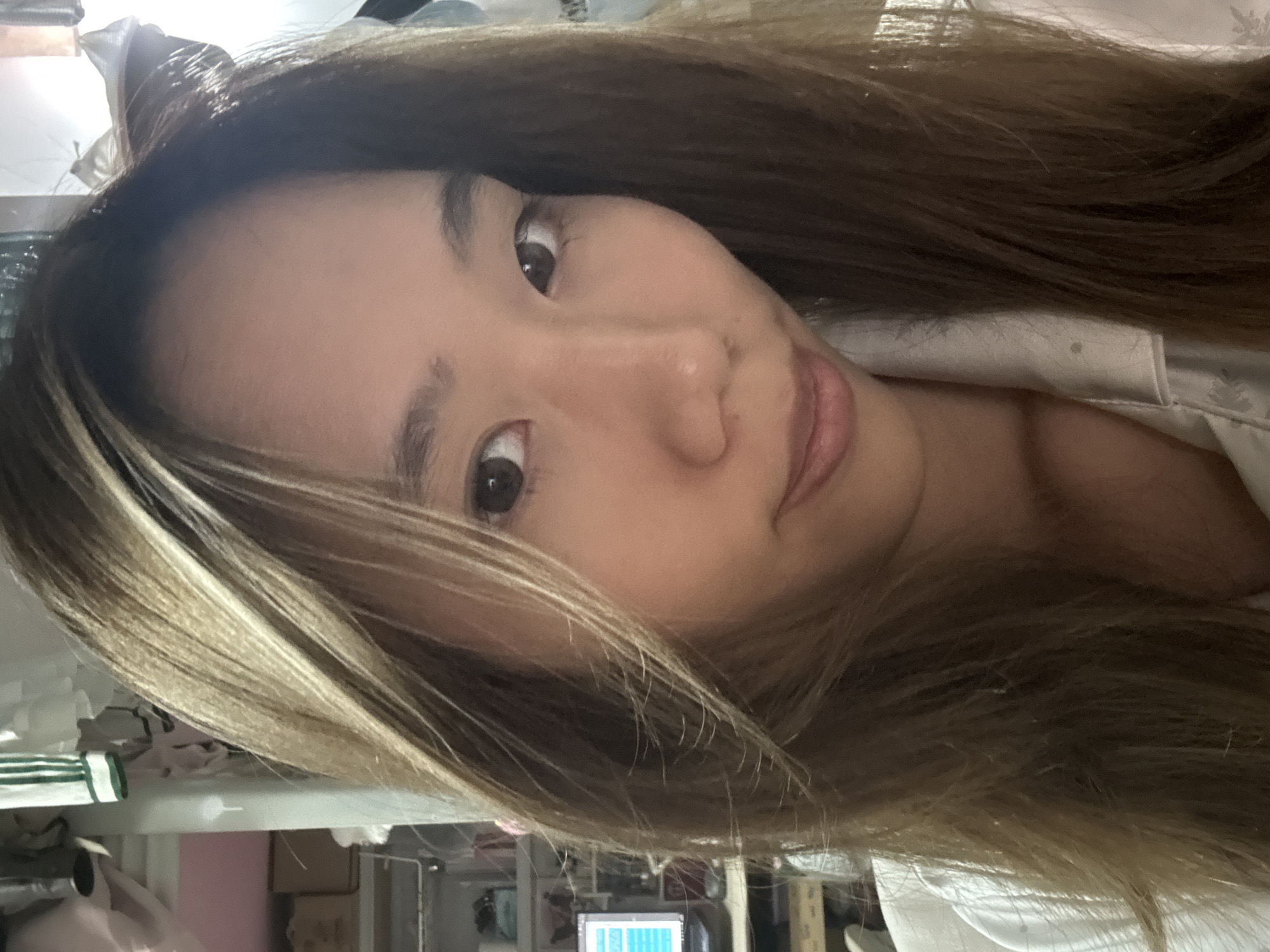 A close-up, slightly angled selfie of a young Asian woman with long, brown hair with blonde highlights is captured indoors. Her facial features include dark eyes, a small nose, and light pink lips. She appears to be wearing a light-colored shirt or pajamas. The background is cluttered with various items including a computer monitor displaying a grid, boxes, and other unidentified objects. The location is unidentifiable as being in Nan'an District, China.