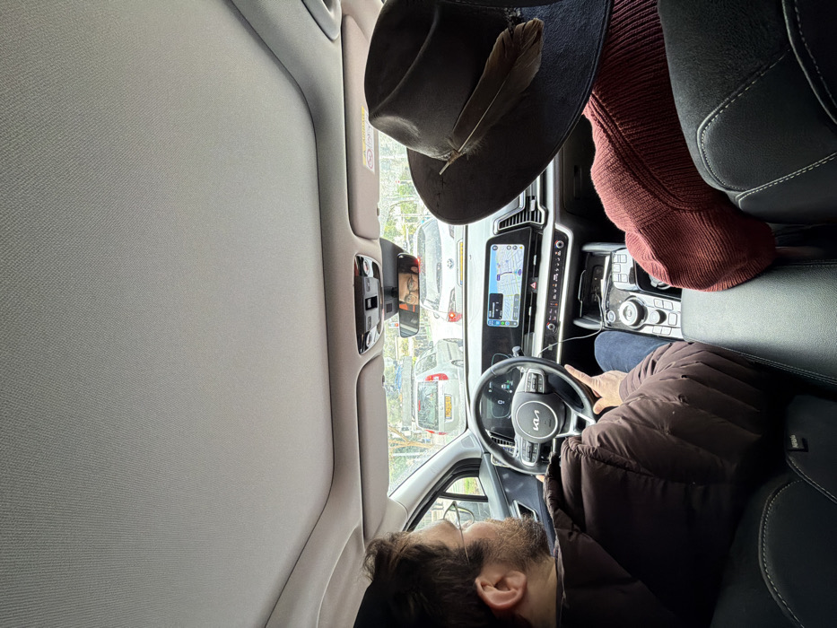The image depicts the interior of a Kia Sportage vehicle, viewed from a rear passenger seat looking forward through the windshield. A male driver is visible on the left, wearing a brown puffer jacket, blue jeans, and glasses. His right hand rests on the steering wheel, which features the "KIA" logo. His face, with short dark hair and a beard, is reflected in the rearview mirror. To the right, a front passenger is partially visible from behind, wearing a dark red ribbed sweater and a dark, wide-brimmed hat adorned with a single brown feather on its right side.
The vehicle's dashboard features a modern, integrated digital display showing both the instrument cluster and a navigation map. The central console includes a rotary gear selector and various control buttons.
Through the windshield, a street scene in an urban environment is visible. The vehicle is in traffic, with a silver hatchback directly in front, bearing an Israeli license plate displaying yellow characters on a blue and yellow background, including the numbers '90-02'. Behind the hatchback, a white sedan is partially visible. Distant trees and buildings suggest a city setting. The scene is located on an urban street in Tel-Aviv, Israel.