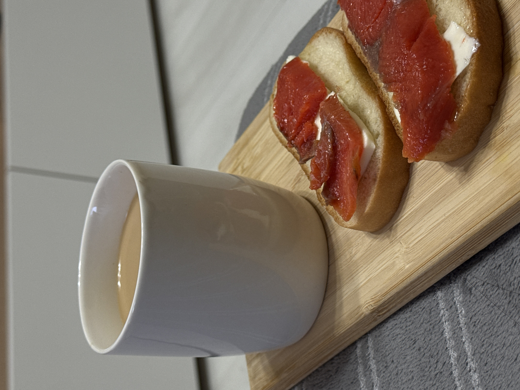 A high-angle shot presents a breakfast scene, featuring a white ceramic mug filled with a beige liquid, possibly coffee or tea with milk, positioned alongside a wooden cutting board on which are two slices of bread topped with a white spread and red salmon. The background consists of a gray cabinet and a striped gray towel. The location is not specified.