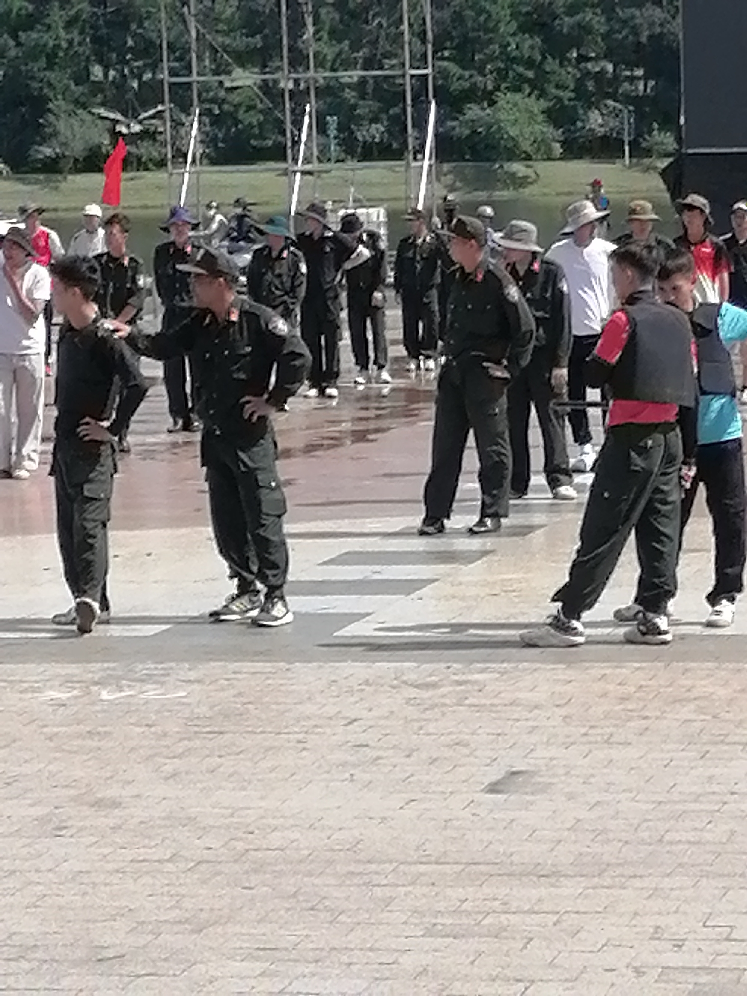 The image depicts a group of individuals, some in uniform, standing on a paved outdoor area. The background suggests an urban or park setting with trees and structures resembling scaffolding. The individuals are dressed in a mix of casual and uniform attire, with some wearing black uniforms and caps. One person in the foreground is wearing a black vest over a red shirt. The ground is paved with interlocking blocks. The presence of a red flag in the background suggests a possible event or ceremony. The overall scene appears to be a gathering or a demonstration in Da Lat, Vietnam, possibly in a public square or recreational area.