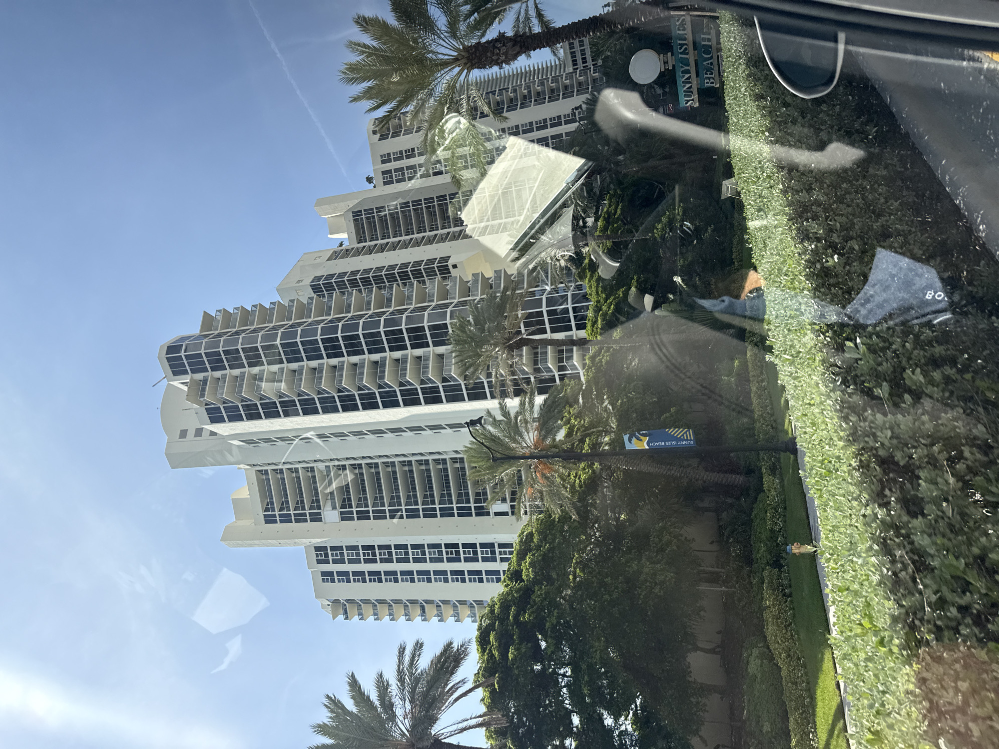 The image shows a sunny day in Sunny Isles Beach, United States of America. A tall, white apartment building with many balconies dominates the background. In the foreground, there are lush green bushes and trees, including palm trees. A sign reading "SUNNY ISLES BEACH" is visible, along with a lamppost. Reflections on the surface of a window obscure parts of the scene. The overall impression is of a residential area near the coast.