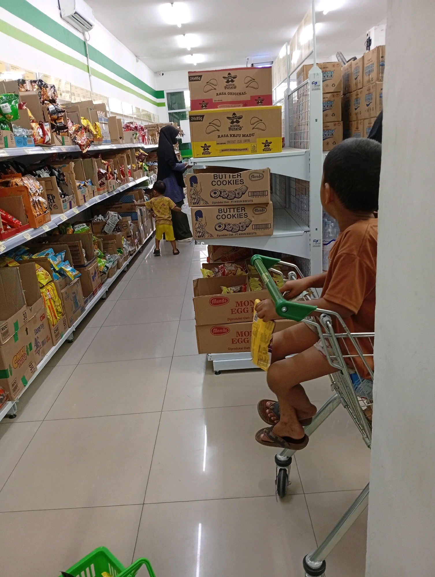In a supermarket aisle in Babakan, Indonesia, a young boy wearing an orange t-shirt and shorts sits in a shopping cart. The cart is pushed by an unseen adult, though a woman wearing a black hijab stands nearby, appearing to select items from a shelf. The aisle is filled with shelves stocked with various products, including boxes of cookies and snacks. The flooring is tiled, and the lighting suggests an indoor environment. The boy is holding a yellow packet and looking towards the products on the shelves.