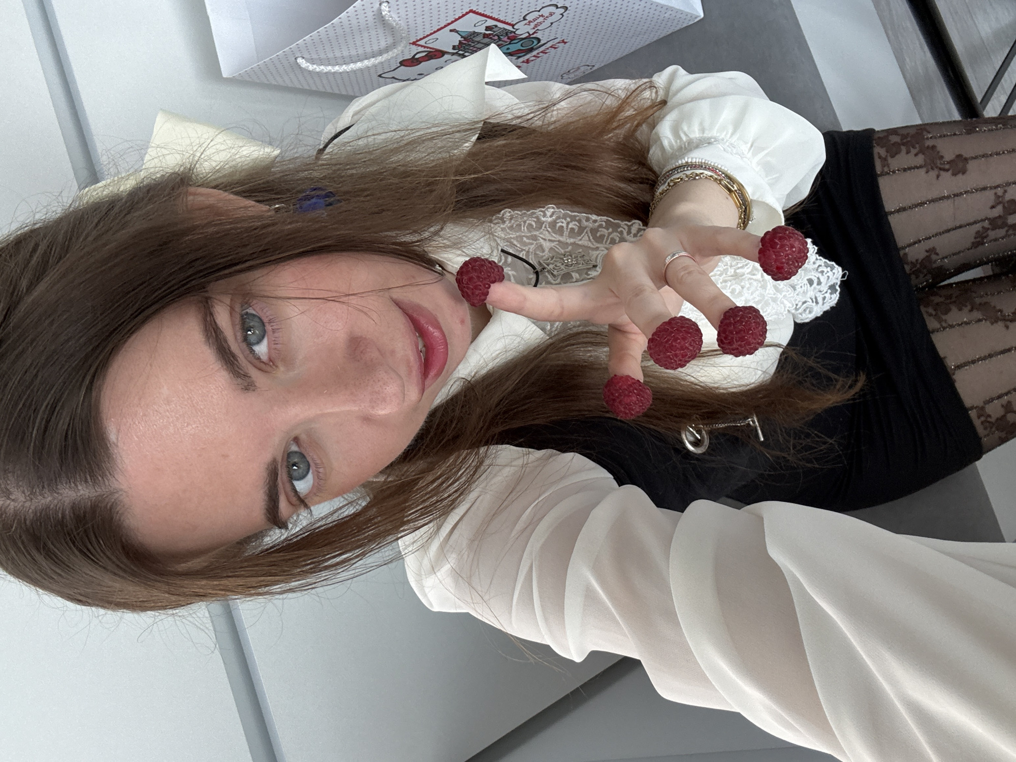A woman with long brown hair and blue eyes is holding four raspberries on her fingers. She is wearing a white ruffled blouse over a black dress, sheer patterned tights, and several bracelets. She also has a ring on her left hand. A white shopping bag with a Hello Kitty design is visible to her right. The background appears to be a modern interior with grey walls. The image is a close-up selfie.
