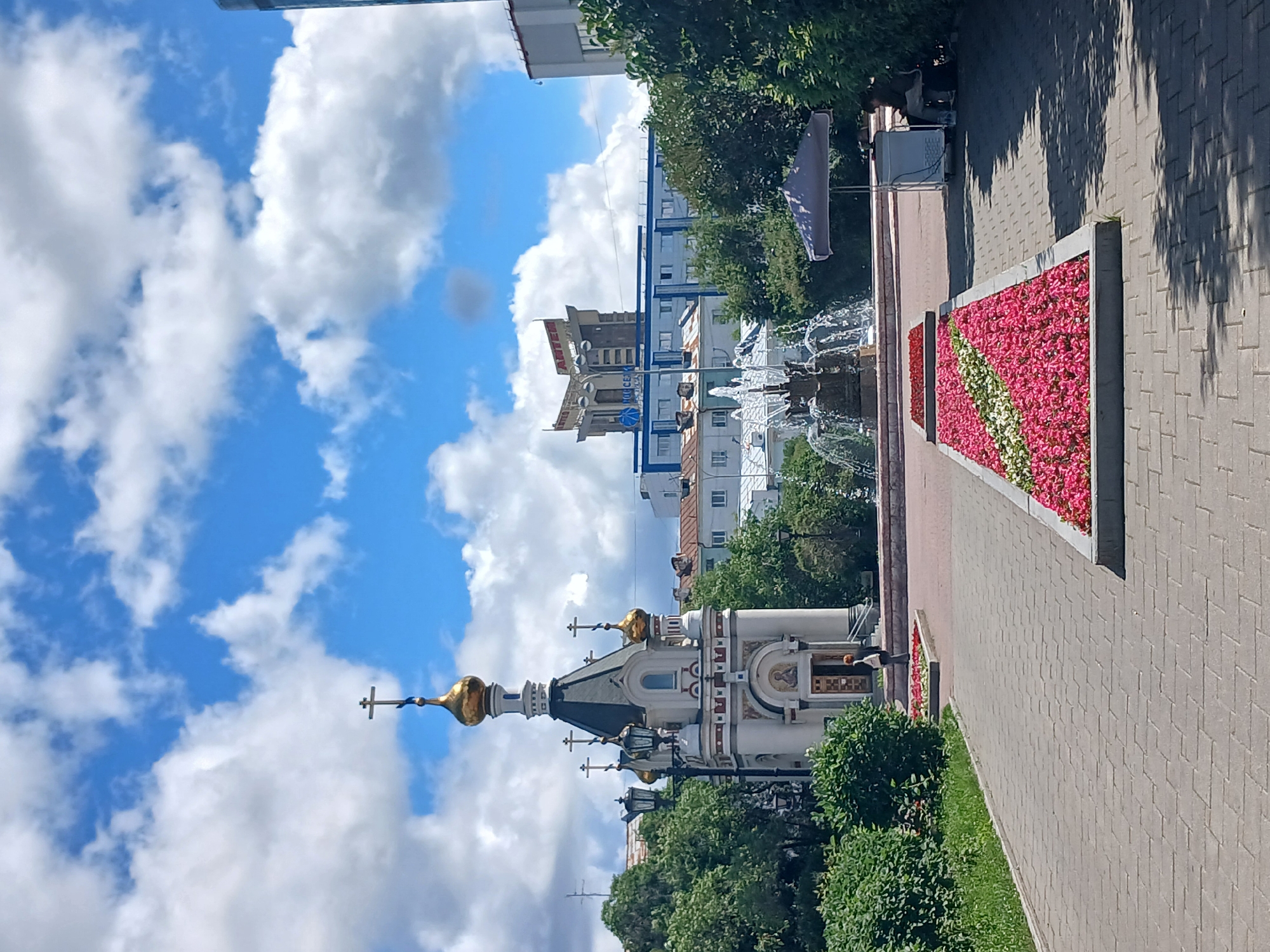 The image shows a sunny day in Yekaterinburg, Russia, with fluffy white clouds against a bright blue sky. In the foreground, a paved walkway leads past manicured flower beds filled with vibrant pink and red flowers. To the left, a small, ornate chapel with golden domes and crosses stands surrounded by lush green trees. In the background, a multi-story building with blue accents and a distinctive angular structure on top is visible. Further to the right, a fountain sprays water into the air. An open-air stall with a grey umbrella offers shade, and a person sits nearby. The scene conveys a sense of peace and urban serenity.