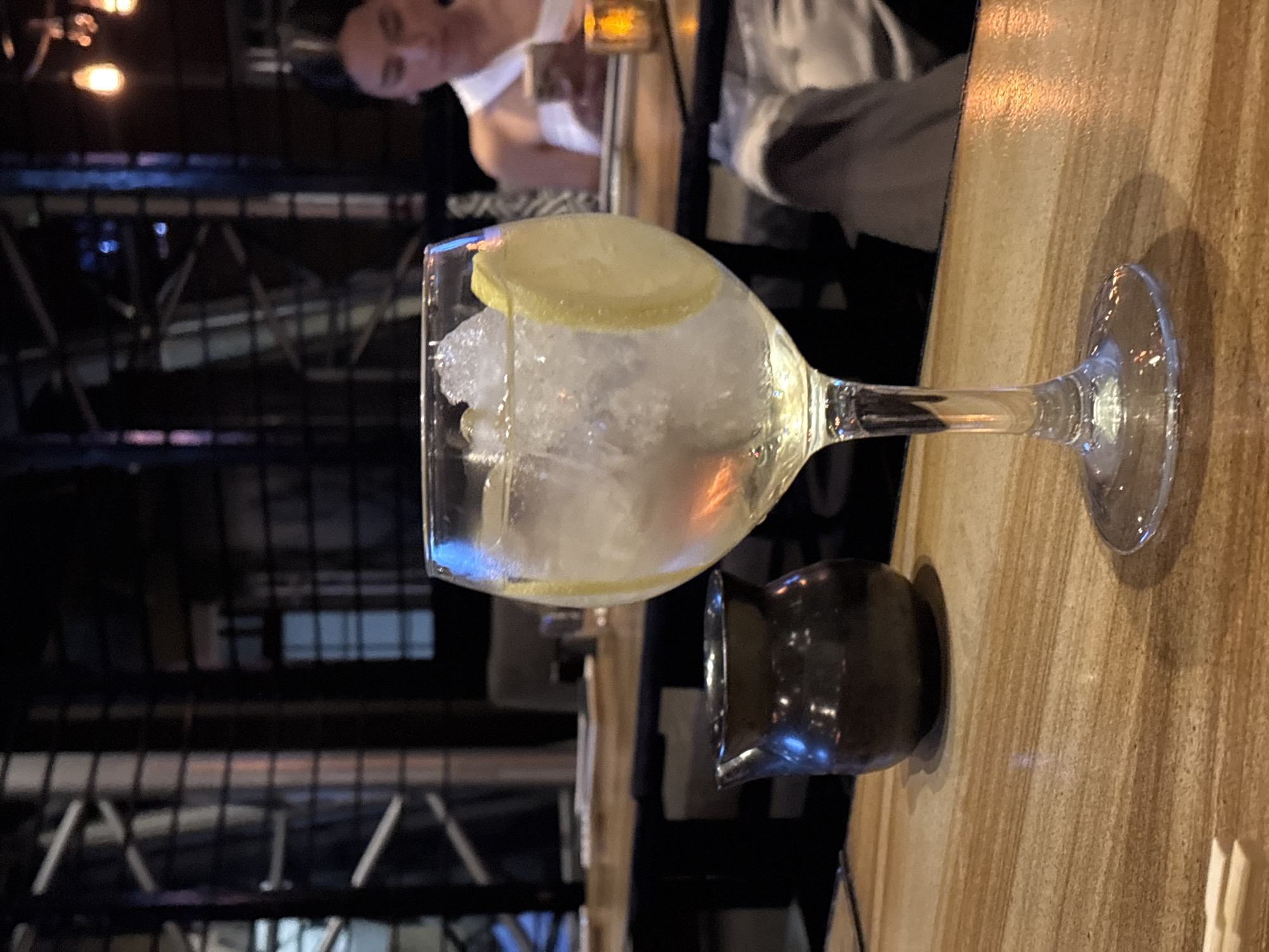 A wine glass filled with ice and a lemon slice sits on a wooden table. Next to it is a small, dark container. In the background, a person is visible. The location is implied to be Puebla, Mexico, although there is no specific information in the image to confirm this. The environment appears to be a bar or restaurant, given the setting and drink.
