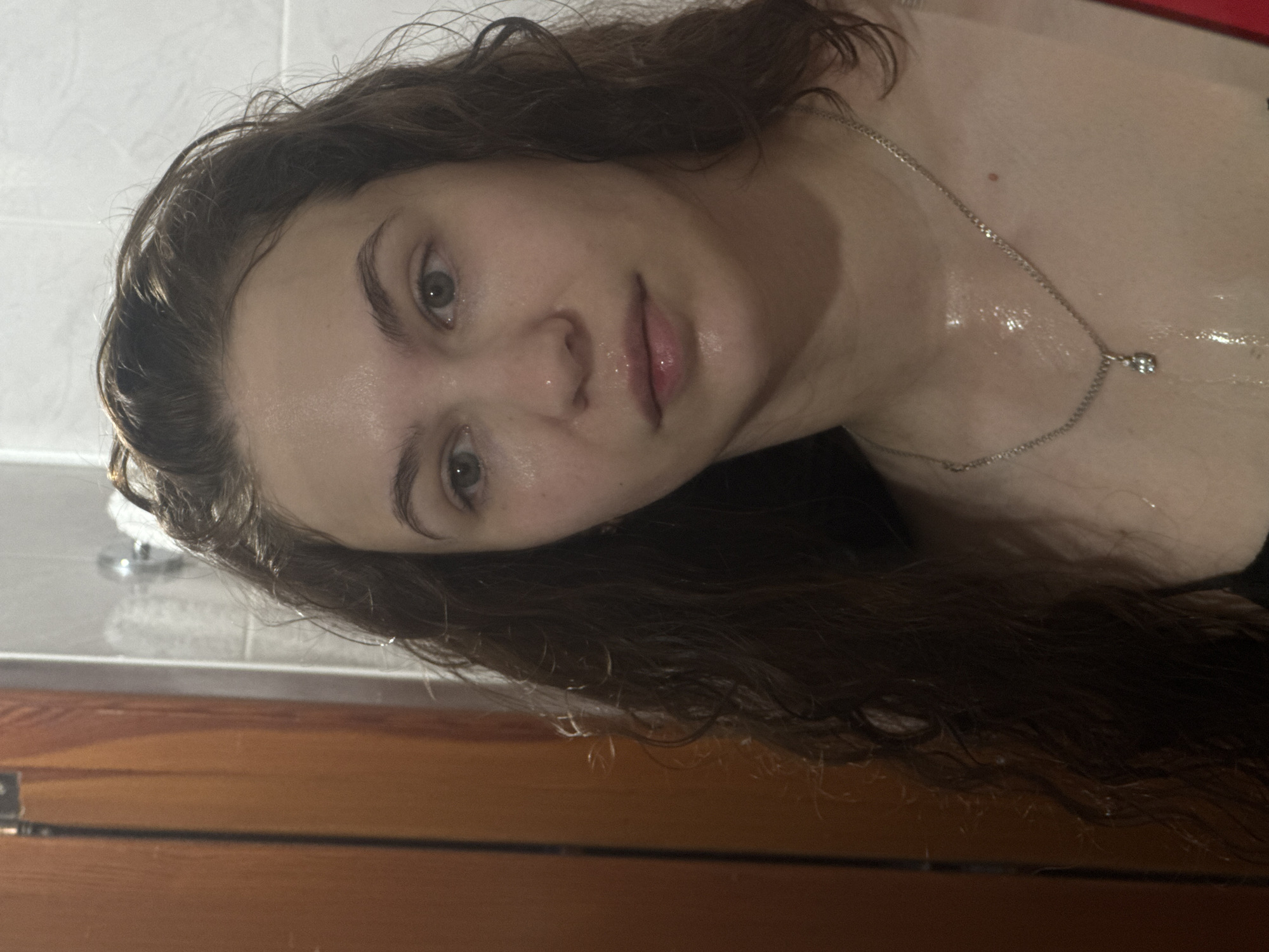 A close-up, portrait-style photograph features a young woman with fair skin and long, wavy, dark brown hair. Her hair and face appear wet, with visible water droplets on her forehead, cheeks, and strands of hair, suggesting she has recently been in water or showered. Her eyes, which are light in color, look directly towards the viewer with a neutral, calm expression. Her lips are slightly parted and appear wet.
She is wearing a thin silver chain necklace with a small, reflective pendant that rests on her collarbone. A dark strap, possibly from a top or swimsuit, is visible on her left shoulder area. The background elements suggest an indoor setting, likely a bathroom. White tiles are visible in the upper part of the image, while a wooden surface, possibly a door or cabinet, occupies the lower portion. The lighting is soft and consistent with indoor artificial light. There is no visible text in the image. The overall impression is an intimate moment captured after being in water, potentially in Chaikovsky, Russia.