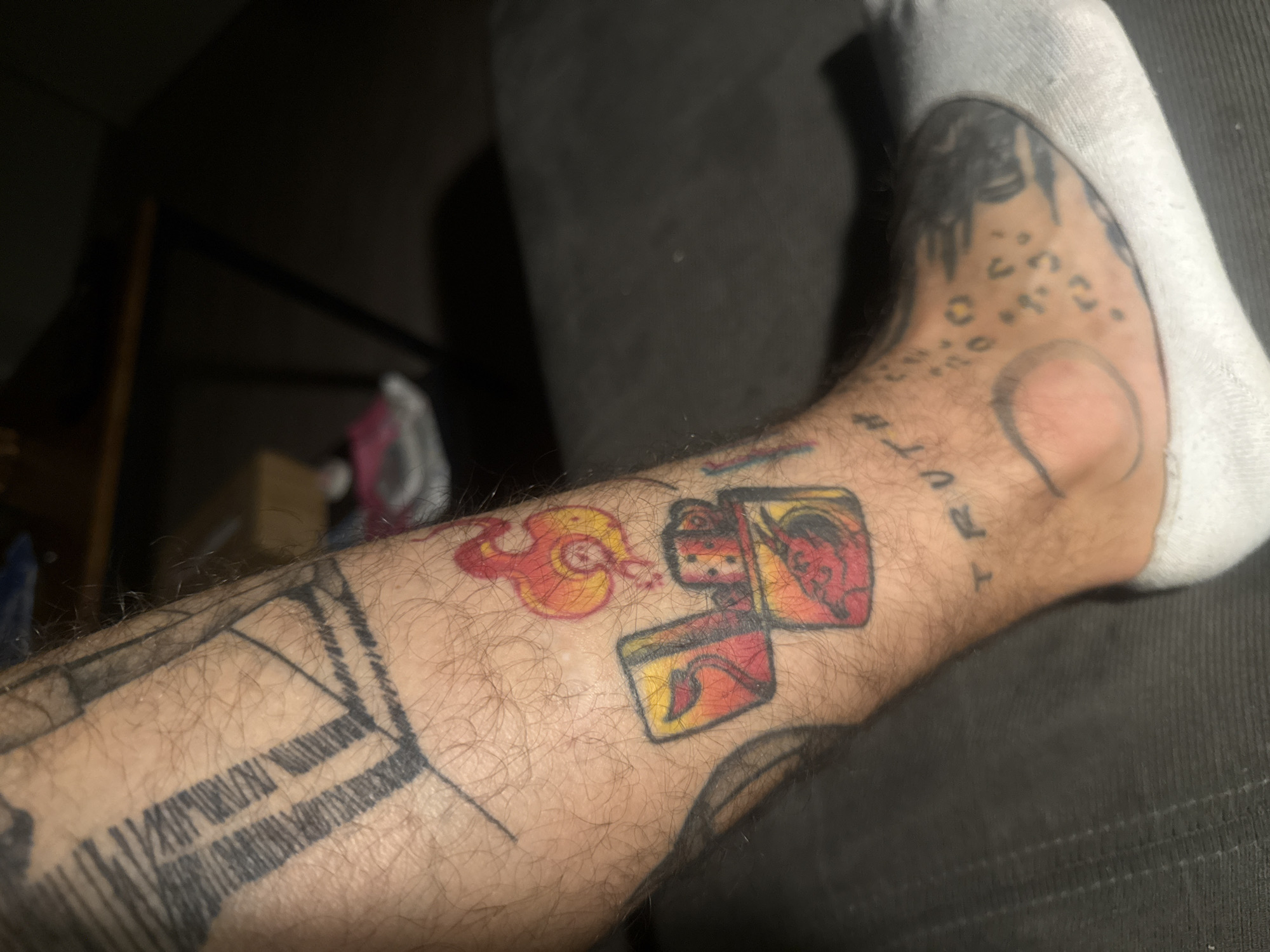 The image shows a person's lower leg with several tattoos. One tattoo depicts a Zippo lighter with an image of a devil on the lighter insert. There's a flame tattooed near the lighter. Other tattoos include a geometric design with parallel lines and what appears to be the word "TRUTH". The person is wearing a white sock. The environment includes part of a dark upholstered chair and a glimpse of a dark-toned room. The scene does not appear to be in Sofia, Bulgaria, as there is no indication of location.