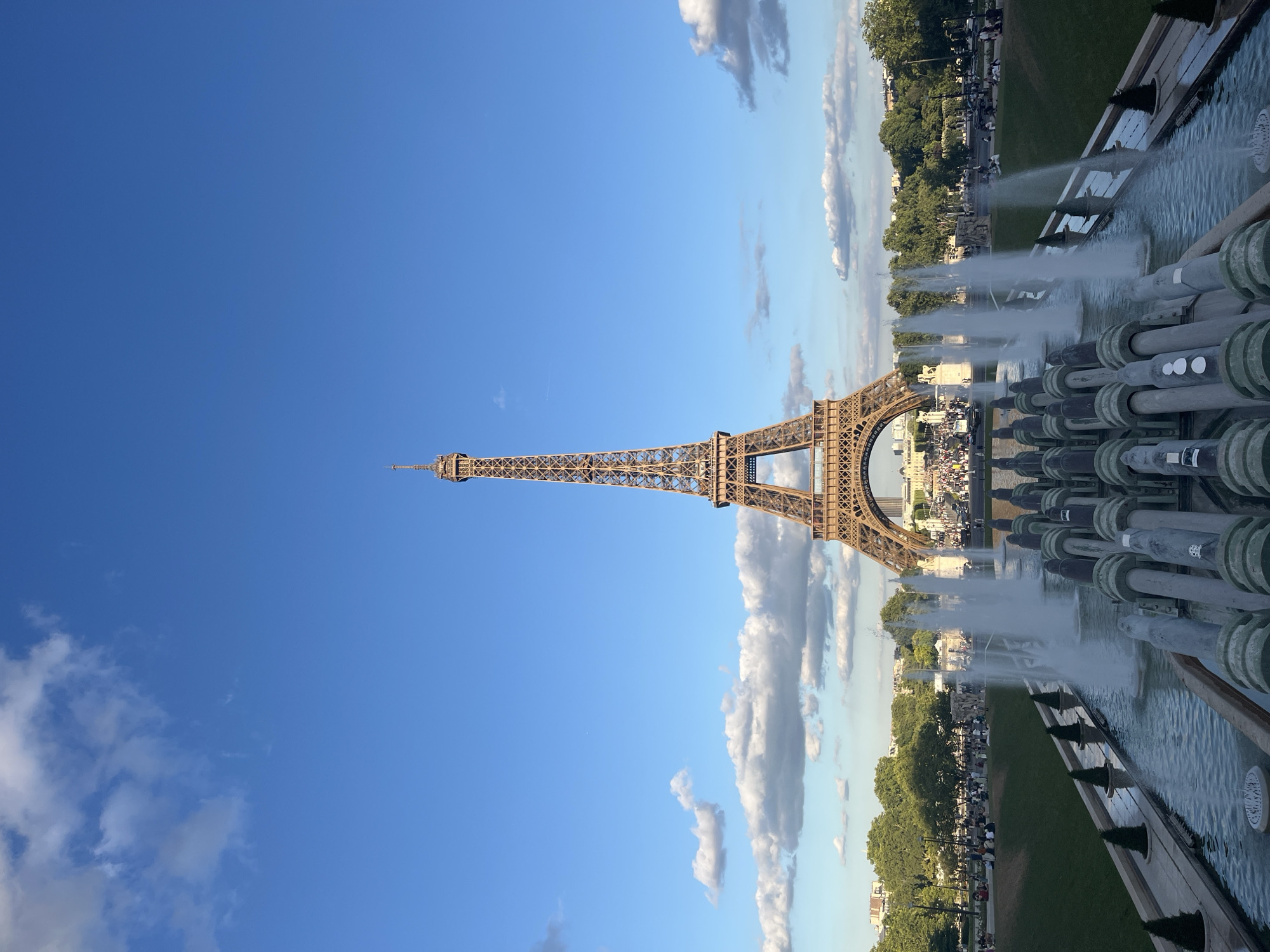 The image showcases the iconic Eiffel Tower in Paris, France, set against a clear blue sky with scattered clouds. The tower, a wrought-iron lattice structure, dominates the upper left portion of the frame, its intricate design visible. In the foreground, a series of large, grey pipes with green accents are part of a fountain. Water sprays upwards from multiple points in the fountain, creating a misty effect.
Beyond the fountain, a lush green lawn stretches out, dotted with trees and people. The people appear to be enjoying a day outdoors, with some gathered near the fountain and others strolling through the park. The scene suggests a pleasant, leisurely atmosphere. The sunlight indicates it is likely daytime, possibly late afternoon given the soft lighting. The presence of the Eiffel Tower and the ornate fountains strongly places the scene in Paris, specifically the Champ de Mars area. No visible text is present in the image.