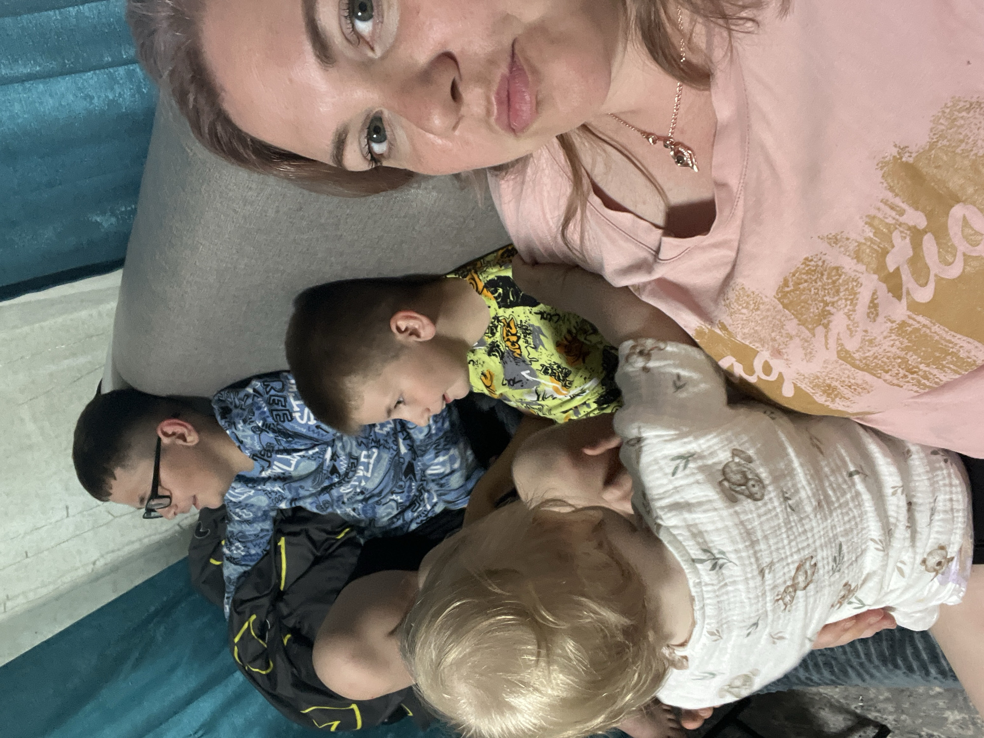 A medium shot photo shows a woman holding a baby on a gray couch. The woman is in the foreground, facing the camera, wearing a pink t-shirt and a necklace. The baby, facing away from the camera, is wearing a white onesie with a print of animals and leaves. To the left of the woman and baby, two young boys are sitting on the couch. The boy in the middle, closer to the woman, is wearing a yellow t-shirt with a black and white graphic print. The boy on the far left is wearing glasses and a blue t-shirt with a camouflage pattern. He is leaning against a black sleeping bag. In the background, a teal curtain is visible. The overall environment appears to be a living room.