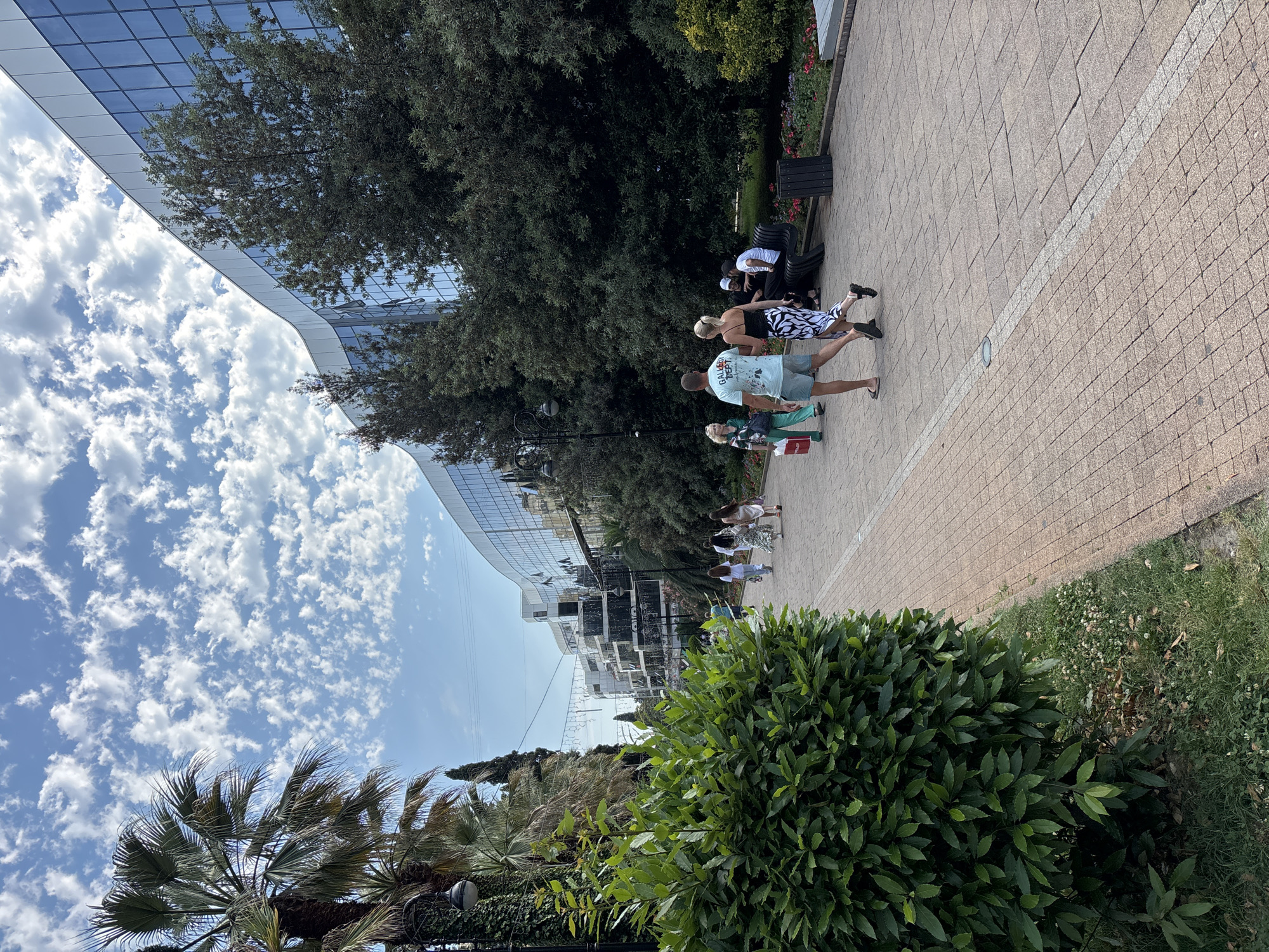 In Sochi, Russia, a paved promenade stretches along a waterfront, flanked by lush green foliage and palm trees. Modern buildings with reflective glass facades rise in the background, their curved structures catching the sunlight. Several people are seen strolling along the promenade. A couple, a man in a light blue t-shirt and shorts and a woman in a black top and a patterned skirt, walk away from the camera. Nearby, a group of individuals are seated on a black park bench, enjoying the outdoors. The sky above is a clear blue, dotted with fluffy white clouds. The scene suggests a pleasant day in a vibrant urban setting.
