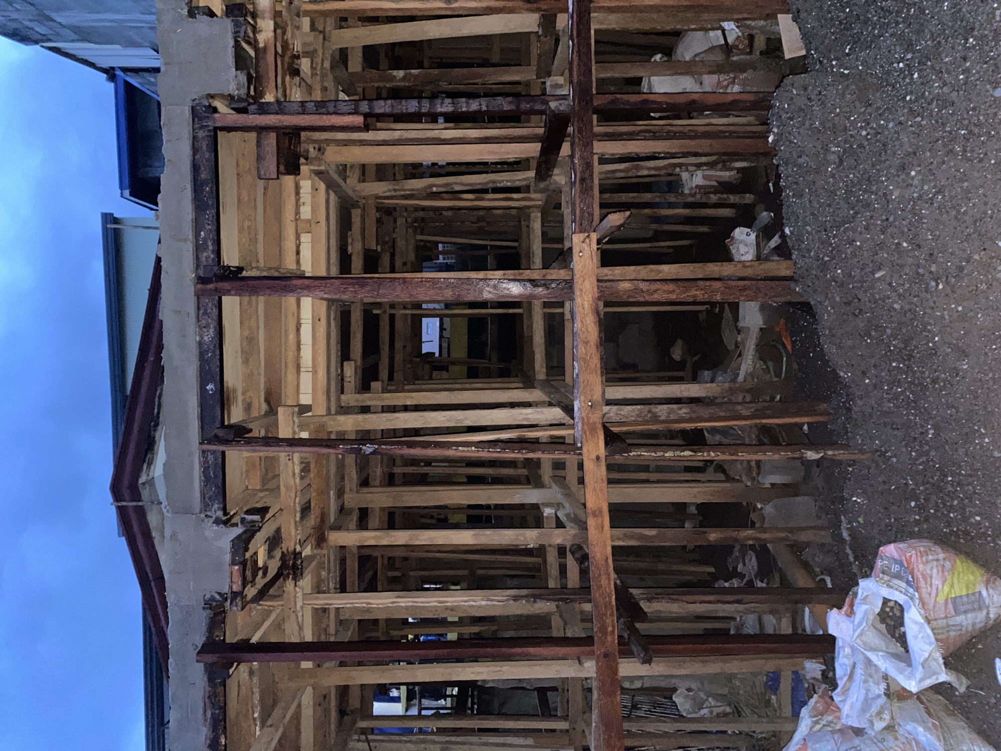 The image depicts the unfinished construction of a building in Davao City, Philippines. The scene is dominated by wooden scaffolding and concrete structures, suggesting an ongoing construction project. In the foreground, several white and red sacks are visible, some of which appear to contain building materials, such as gravel or sand. The ground is covered with loose gravel and dirt. The sky is overcast with a twilight hue. There are no people clearly visible in the frame. The overall impression is one of an industrial or construction site.