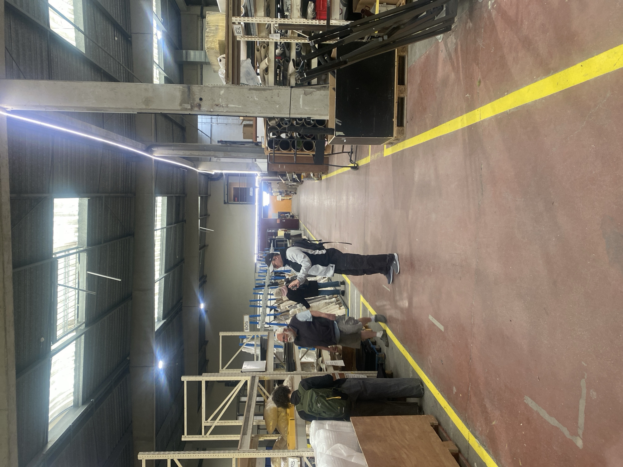 An indoor scene depicts a spacious industrial warehouse or workshop, likely in Montreuil, France. The primary subjects are four individuals, all appearing to be adult males, engaged in observing or inspecting materials stored on tall metal shelving units.
The floor is a reddish-brown concrete, marked with a prominent yellow safety line that delineates a walkway down the center of the facility. Along both sides of this aisle, multiple industrial-style metal racks are filled with various raw materials and components, including rolls of fabric or insulation, metal bars, and wooden planks. Visible text on the shelves includes "METAL", "BOIS" (French for wood), and partially visible "LUX" and "RS". Wooden pallets, some marked with "EUR", are also present.
The ceiling is high, featuring exposed concrete beams and corrugated metal panels. Bright strip lighting runs along the ceiling, complementing the natural light streaming in through large, high-set windows.
One person, wearing a black and white jacket, dark pants, and a cap, with a backpack, stands in the foreground, looking downwards. Further down the aisle, another individual with a white beard is seen in a dark vest and shorts, also appearing focused on items on the shelves. A third person, with dark curly hair and a green backpack, stands with their back to the viewer, near the shelves. A fourth person is partially visible behind the first individual. The activity suggests an inspection, inventory, or a guided tour of the facility. The lighting indicates it is daytime.