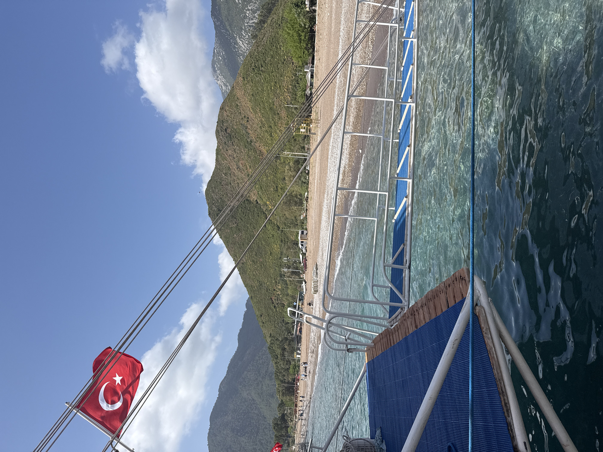 The image captures a view from a boat near the shoreline in Kumluca, Turkey. A sandy beach runs parallel to the mountainous terrain visible in the background. The water near the boat is clear and turquoise. On the boat itself, a Turkish flag is attached to one of the ropes. The boat also has a ladder with blue steps leading down to the water. The scene is set under a blue sky with scattered clouds. The beach is lined with trees and other structures. People are visible on the beach.