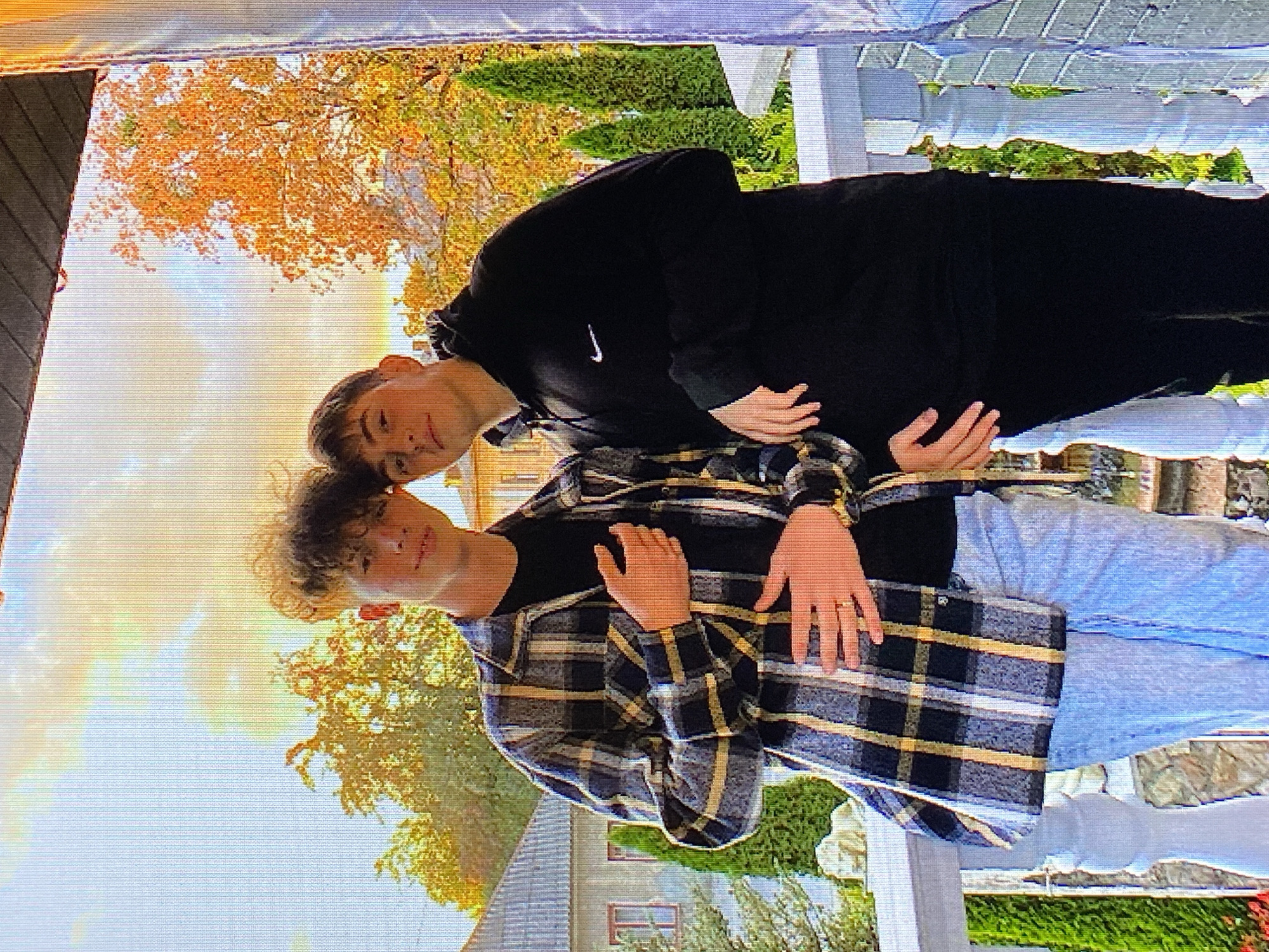 Two young men are posing for a photo on a balcony with a white railing. The man on the left is wearing a plaid shirt over a black t-shirt and light-colored jeans. He has his left hand on his chest and his right hand on his hip. The man on the right is wearing a black hoodie with a Nike logo and black pants. He has his right hand on his hip. Behind them, there are trees with orange and yellow leaves, a building with a red-tiled roof, and green cypress trees. The sky is partly cloudy with a warm, orange glow. The image appears to have been taken in Poznan, Poland, as indicated by the architecture and foliage.