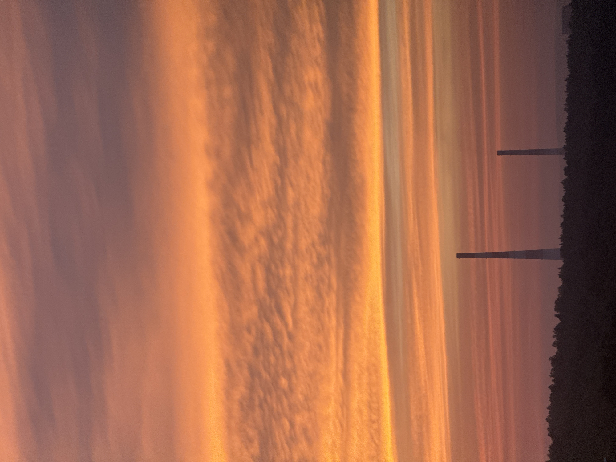 The image depicts a silhouette of a treeline against a gradient sky of orange and purple hues at dusk. Two smokestacks rise above the treeline, one taller and to the left, the other shorter and to the right. The sky is filled with horizontal bands of clouds, reflecting the warm tones of the setting sun. The scene is devoid of human activity or immediate discernible interaction, presenting a stark, industrial landscape under a dramatic sky. The context of Markova, Russia, suggests a potentially remote or industrial area.