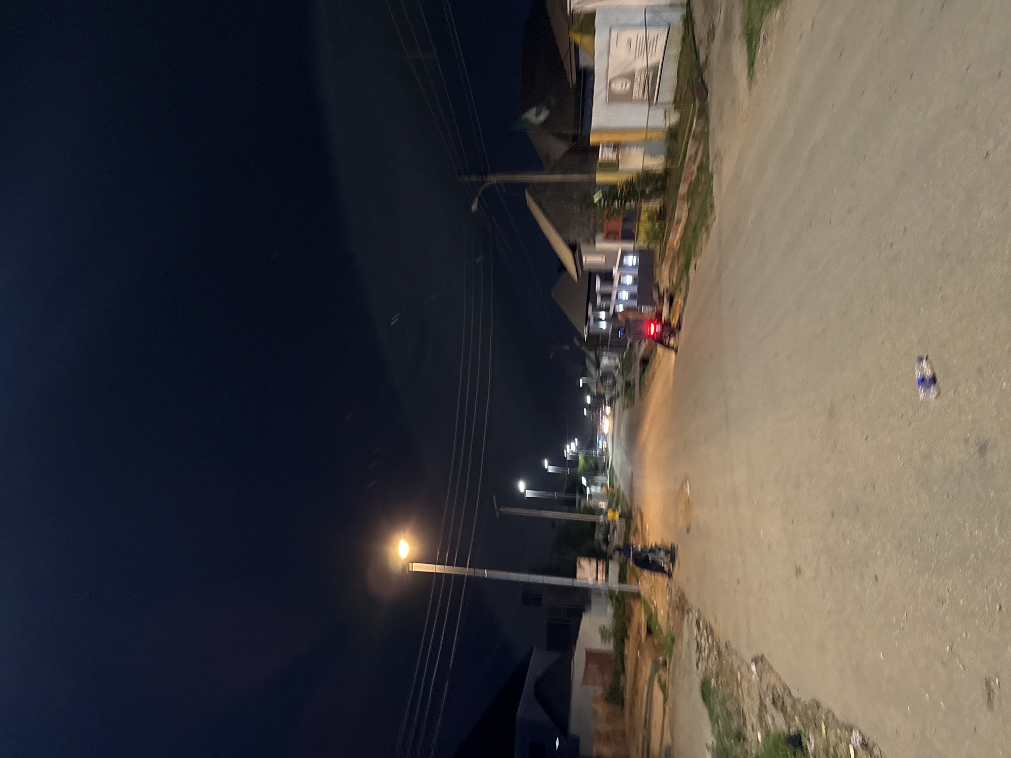 A nighttime, eye-level, medium shot captures a paved street in a residential area of Abuja, Nigeria, illuminated by streetlights and the distant glow of vehicle headlights. The sky is a deep, dark blue, with faint streaks of light suggesting movement or long exposure. On the left side of the street, a motorcycle is parked on the unpaved shoulder, with its rider seated. Further down the street, another motorcycle is visible, with its red taillight illuminated, and a person is standing near it. On the right side of the street, several buildings with peaked roofs line the road. A white fence runs along the property line, with a sign mounted on it. The road itself is mostly empty, with a few scattered pieces of litter on the ground.
