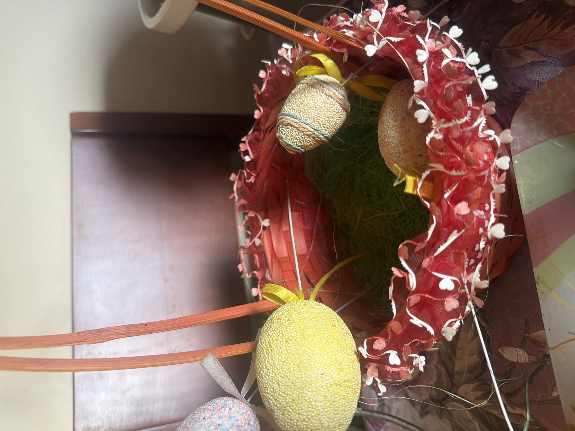 A close-up shot shows an Easter decoration. The main element is a red, bowl-shaped container with a white floral trim, filled with green shredded material resembling grass. Inside this container, two decorated Easter eggs with ribbons are visible. Another Easter egg, colored yellow with a matching ribbon, hangs outside the container, along with a fourth egg that is multicolored. Orange decorative sticks are also part of the arrangement. The background includes a portion of a brown wooden structure to the left, a white object above the decoration, and a surface with a textured pattern.
