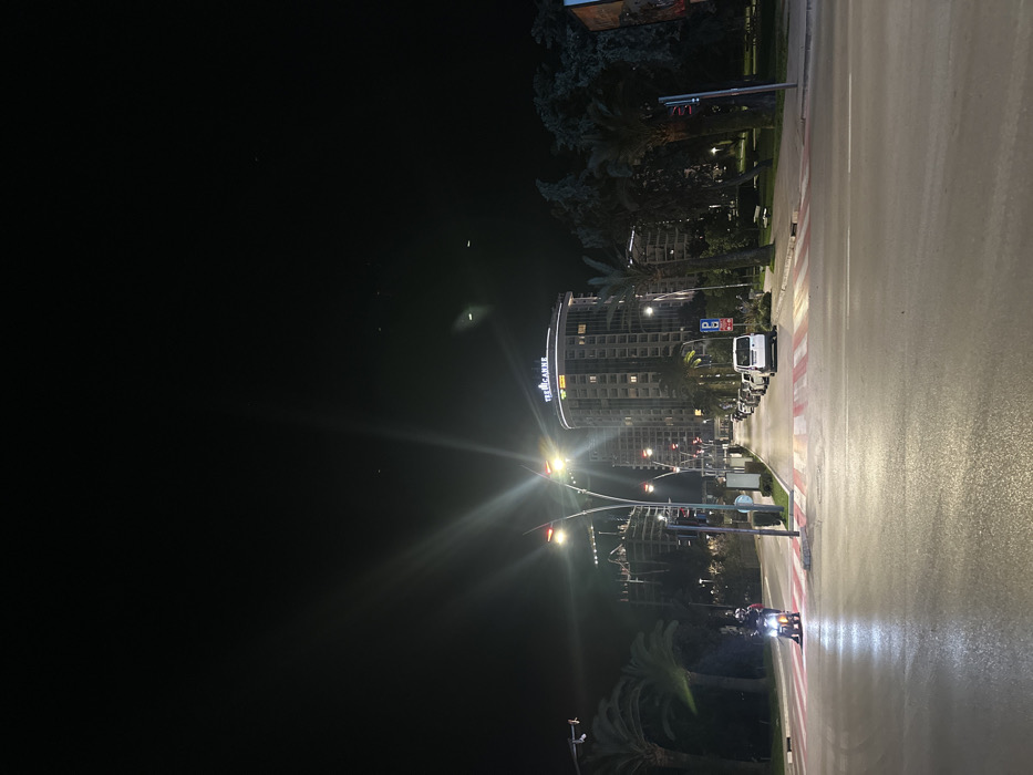The image presents a nighttime street view in Budva, Montenegro, centered on a multi-lane asphalt road with a prominent red and white striped pedestrian crossing. A single individual wearing a helmet is seated on a scooter with an illuminated headlight, positioned on the left side of the pedestrian crossing. The road surface exhibits reflections from bright artificial streetlights, which feature curved fixtures and are casting lens flares. Along the left side of the street, palm trees line a median, behind which an unfinished high-rise building with visible construction elements is illuminated. On the right side of the road, several white vehicles are parked adjacent to a sidewalk, marked by a blue 'P' parking sign. Beyond the sidewalk, a green space with additional palm trees, dense foliage, and a park bench is visible, alongside a red traffic light pole. In the background, the multi-story, curved "TRE CANNE" building is brightly illuminated with its name sign prominently displayed, accompanied by another illuminated tall building to its left. The sky is dark and clear, consistent with night conditions.
