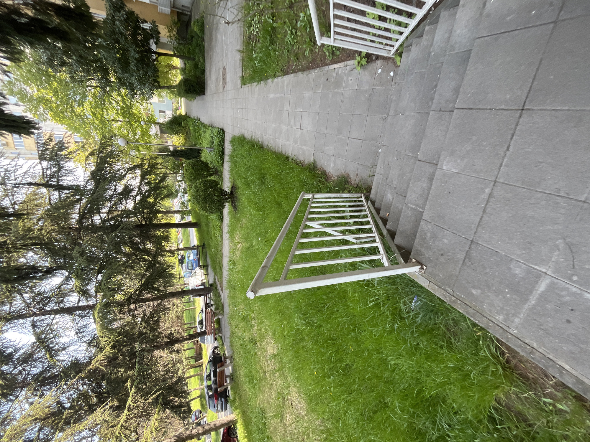 The photograph, taken in Burgas, Bulgaria, presents a ground-level perspective of an outdoor urban environment. A paved walkway extends into the distance, flanked by green grass and trees on the left. A set of gray concrete steps leads upwards from the right foreground. A white metal railing is positioned on the right side of the steps and also lies partially on the grass adjacent to the steps, appearing detached or removed. Cars and park benches are visible through the trees to the left, suggesting a street-side park or residential area. The buildings on the left side of the road are apartment buildings. The time of day appears to be daylight, with even lighting throughout the scene. No people are visible in the image.