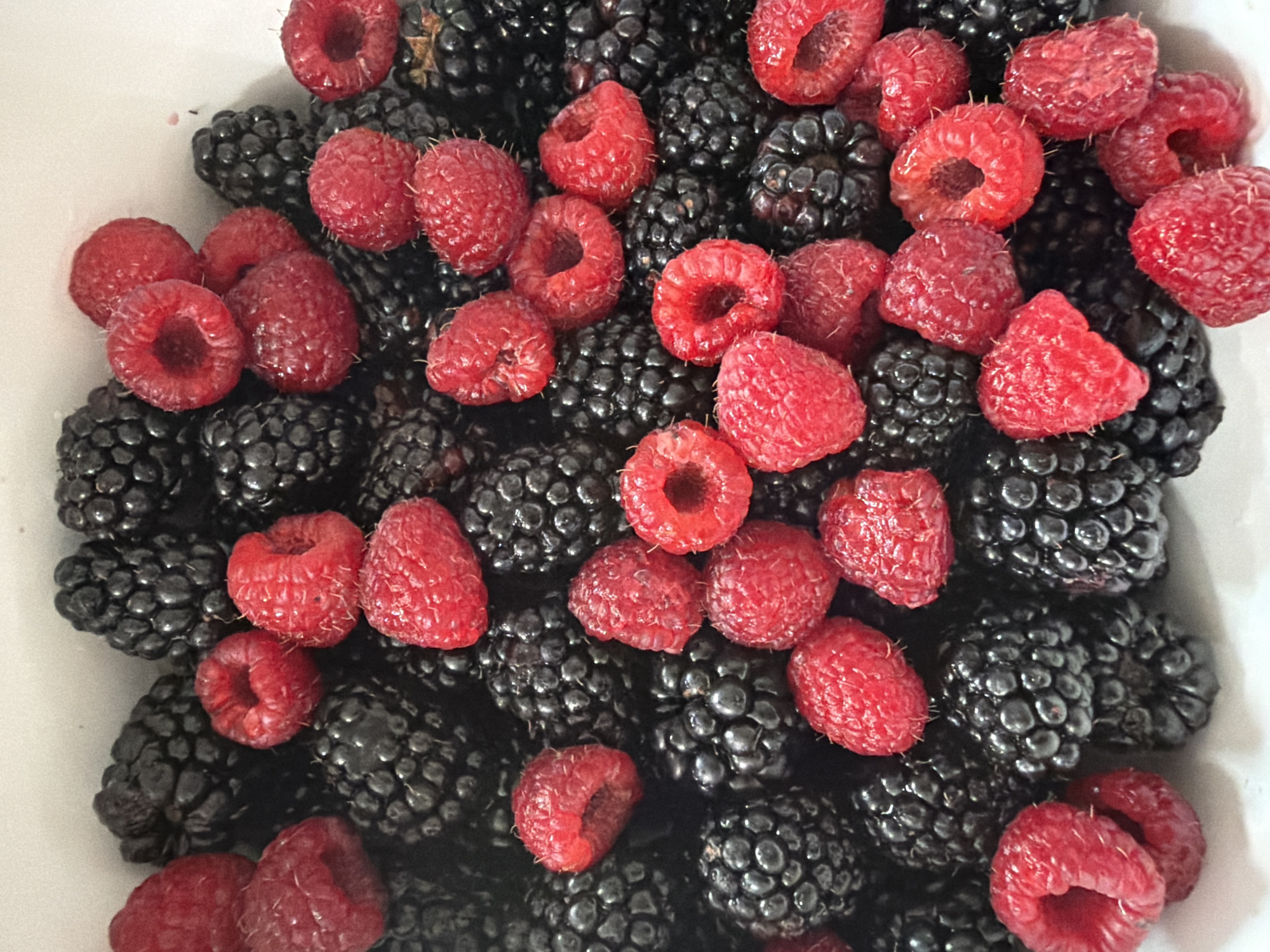 A close-up overhead shot shows a white bowl filled with fresh blackberries and raspberries. The berries are densely packed and appear to be ripe. The lighting is bright, highlighting the textures and colors of the fruit. There are no people or other objects visible in the frame. The location is unknown.
