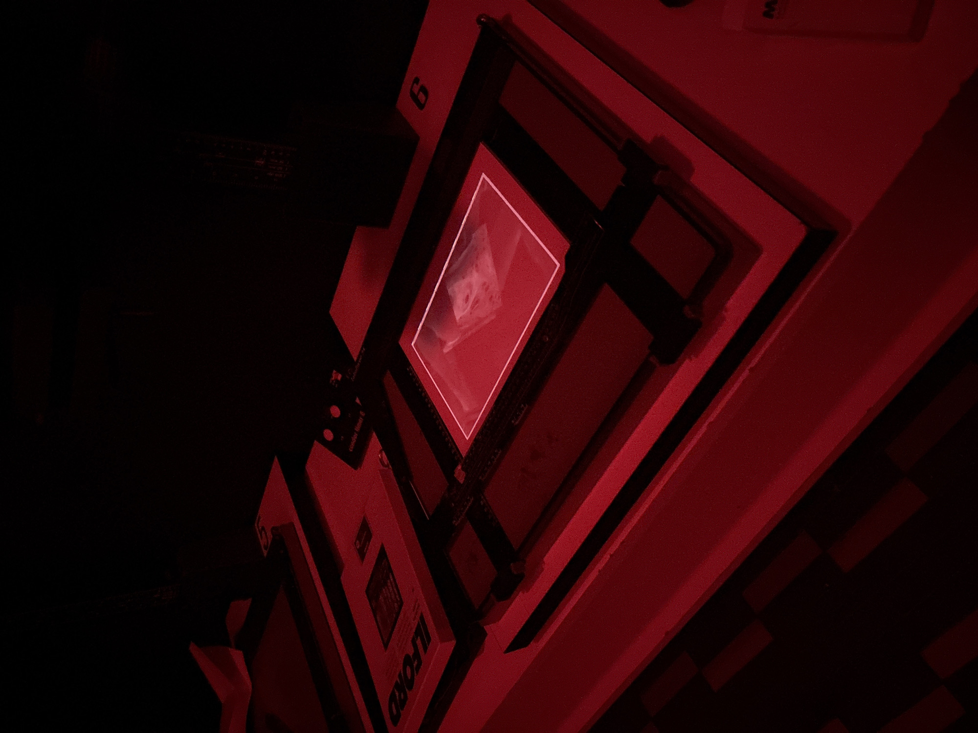 The image shows a darkroom lit with red light. A photographic enlarger sits on a table. Underneath the enlarger, a negative carrier holds a black and white negative. The word "ILFORD" is partially visible on a box of photographic paper. The image is in Milan, Italy.