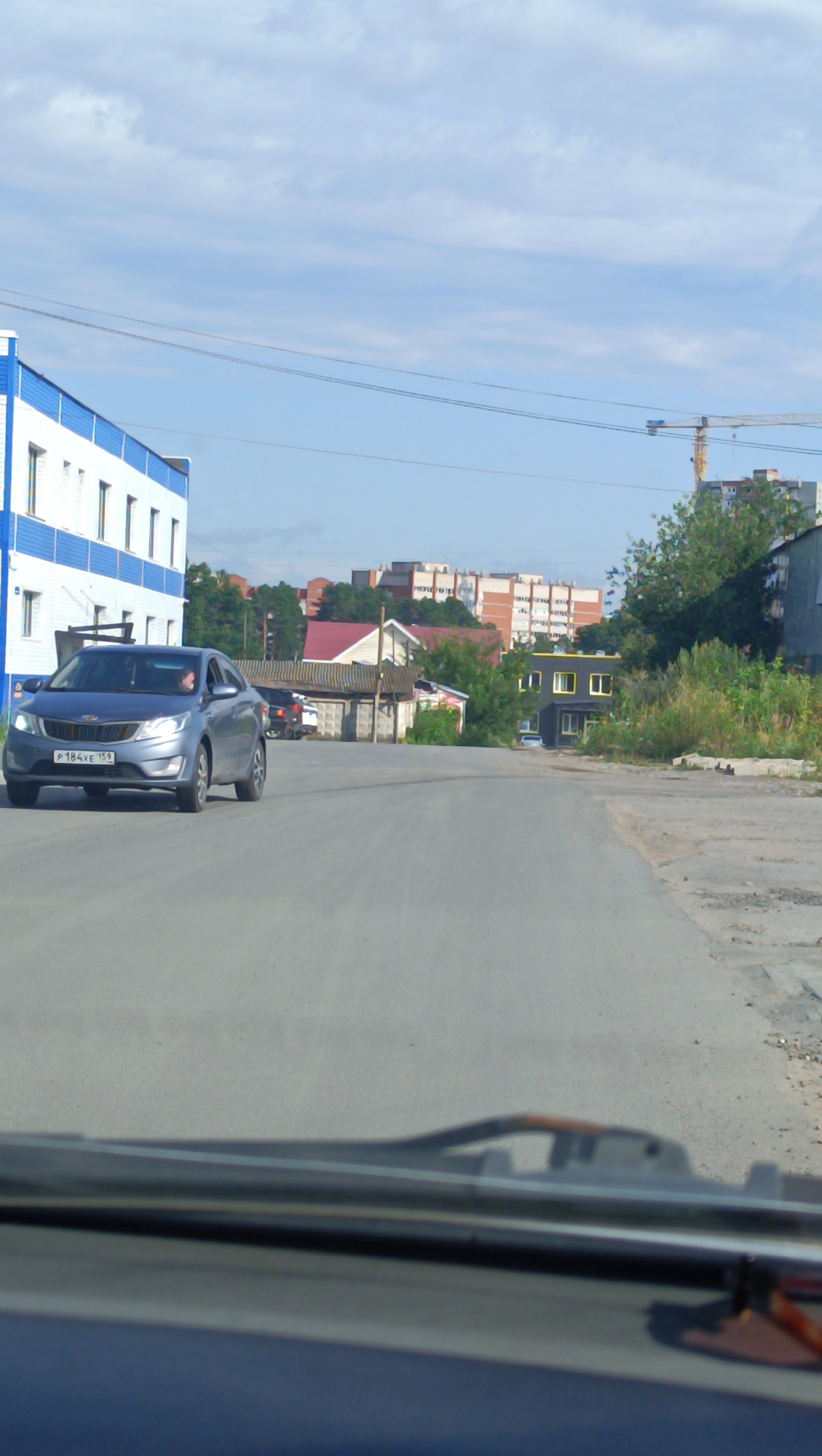 A blue sedan is driving down a paved road in a suburban or industrial area of Chaikovsky, Russia. The car has a license plate that reads "P 184XE 159". To the left of the road is a two-story building with white and blue siding, possibly an industrial or commercial structure. In the background, there are several residential buildings, some of which appear to be under construction, as indicated by a crane. Trees and vegetation are scattered throughout the scene, and the sky is partly cloudy. The viewpoint is from inside another vehicle, looking out through the windshield.