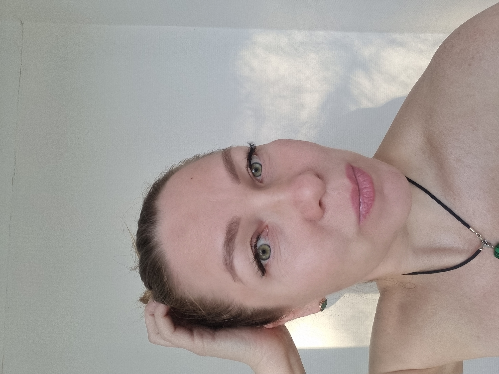 A pale-skinned woman with green eyes is posed against a white wall, tilted to her left. Her hair is pulled back, revealing a green earring. She is wearing light pink lipstick. Her left hand is touching her head near her ear. A black cord necklace with green beads is visible. The background includes subtle shadows. The location is not discernible as Kemul, Russia based on visual information.