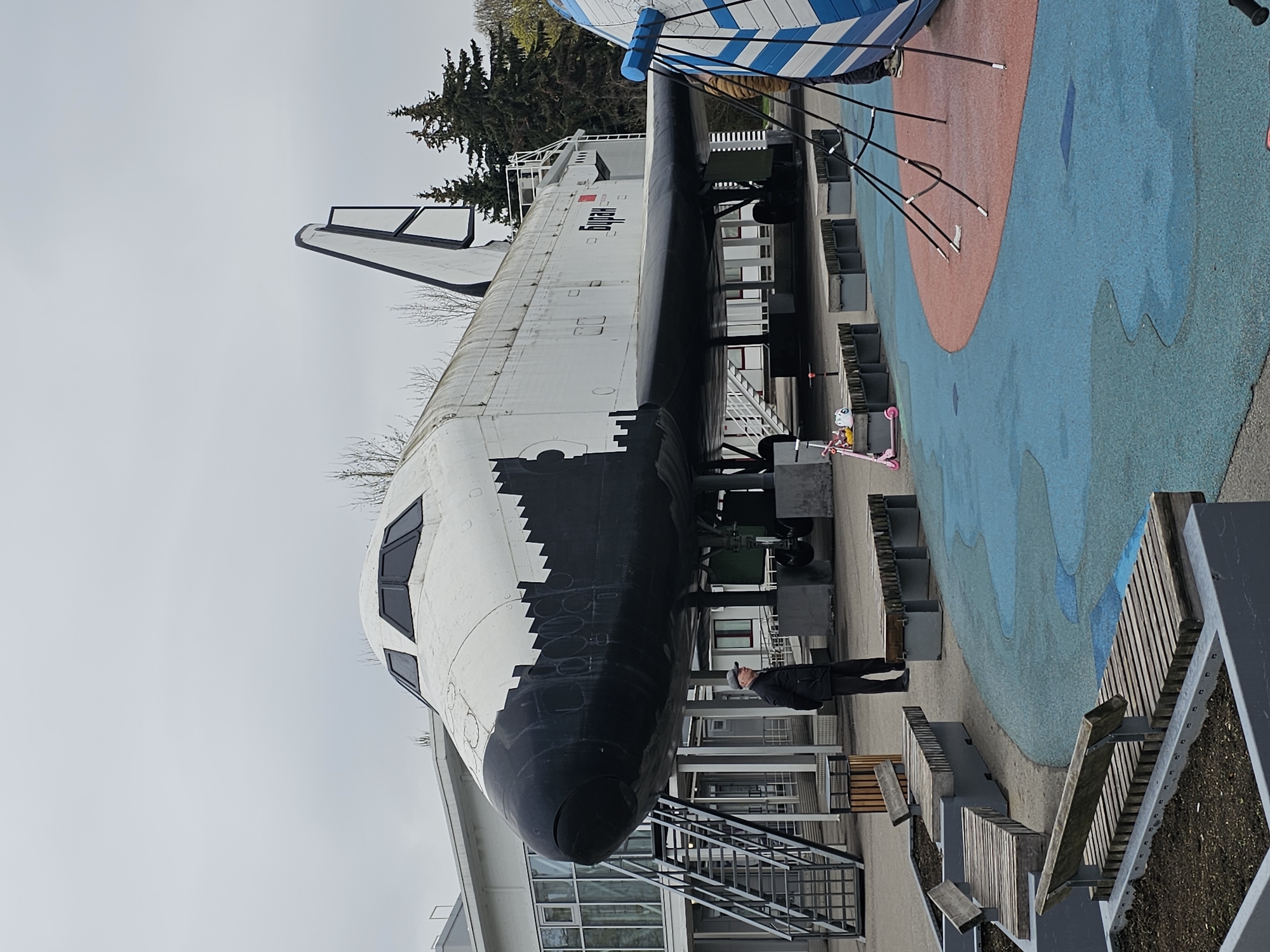 The image displays a large, full-scale Buran-class space shuttle, presented as an outdoor exhibit in what appears to be a public park or museum in Moscow, Russia. The shuttle is predominantly white with black thermal tiles on its nose and underside, featuring a distinctive tail fin. The word "БУРАН" (Buran), written in red Cyrillic lettering, is clearly visible on the side of its fuselage.
The enormous spacecraft is elevated on grey support structures. A lone individual, dressed in a dark jacket and cap, stands beneath the nose of the shuttle, looking up in observation, providing a sense of scale to the massive vehicle. In the foreground and to the right, the ground is covered with a vibrant, patterned surface featuring large swaths of blue and reddish-orange, resembling a recreational or decorative pavement. Modern, slatted wooden benches with grey metal frames are arranged around the display area, suggesting a visitor-friendly environment.
In the background, evergreen trees are visible, and a modern building with large windows can be partially seen behind the shuttle. The sky is overcast and grey, indicating a cloudy daytime setting. The scene captures a moment of contemplation or interest as the person observes this significant piece of space history.