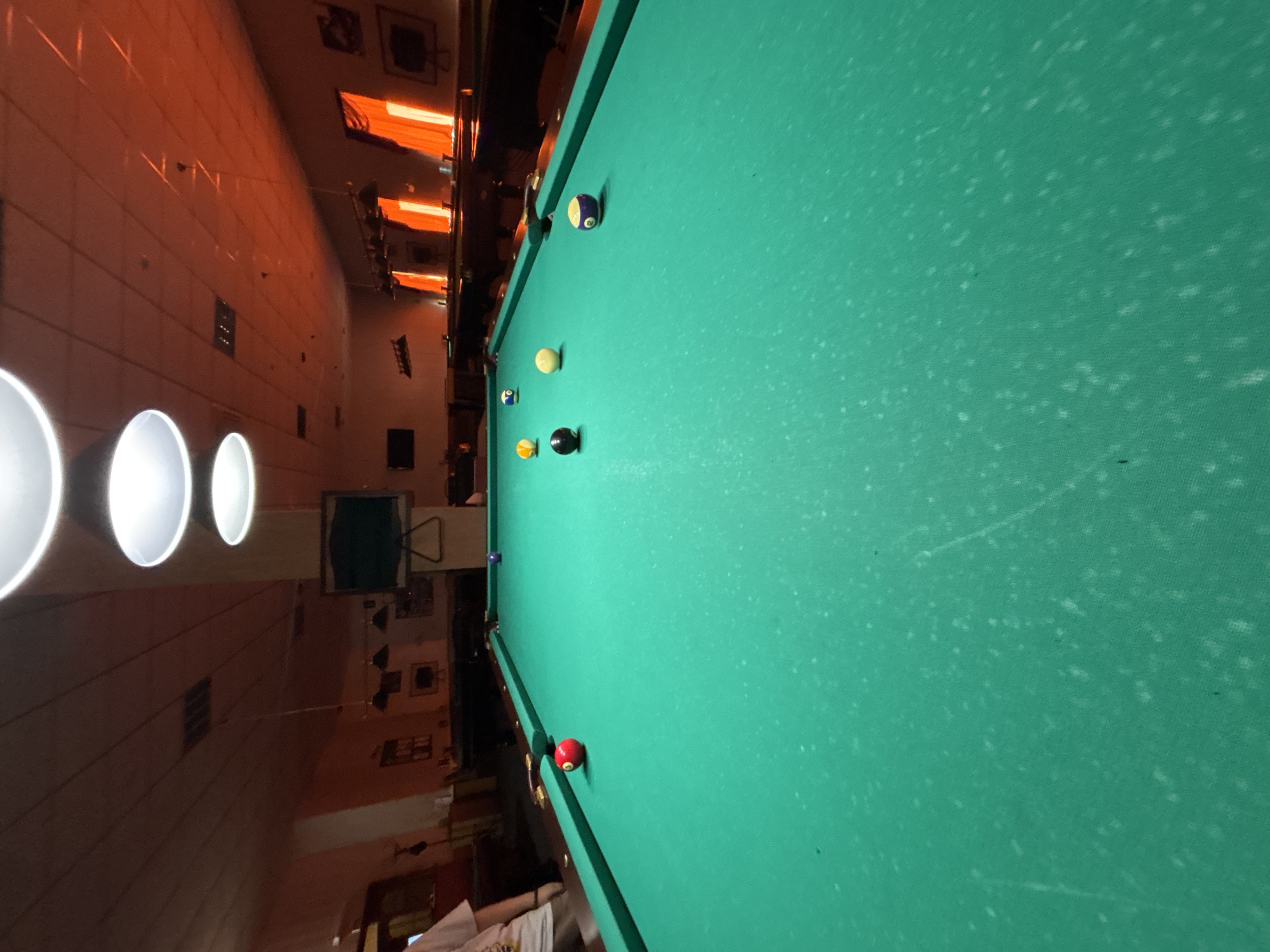 The image depicts a billiard table in a room with orange-toned walls. The table is green with several billiard balls scattered across it. The location is potentially Chaikovsky, Russia, based on prior context, though the image itself provides no information to confirm this location. There are no people visible in the main view of the billiard table. The environment includes overhead lighting and glimpses of furniture and decor.