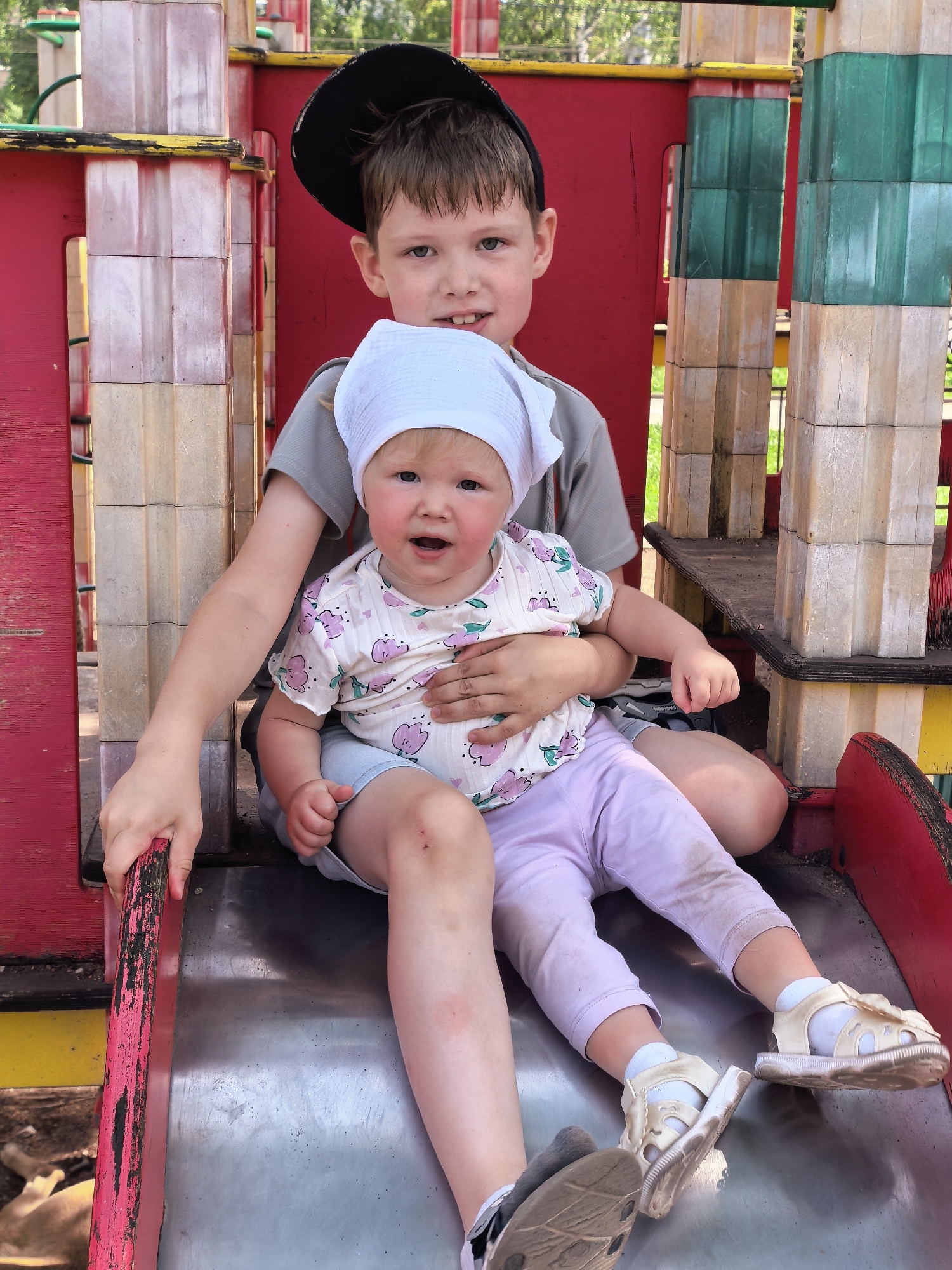 Two children are on a metal slide at a playground. The older child, a boy wearing a black cap and a grey t-shirt, is seated behind a younger child, a girl wearing a white headscarf and a patterned shirt with pink flowers. The girl is being held by the boy and appears to be looking at the camera with an open mouth. Both children are on the slide, with their legs extended. The playground structure is made of red and green painted metal and wood. The ground beneath the slide appears to be dirt and leaves. The image was taken in the city of Sterlitamak, Russia.