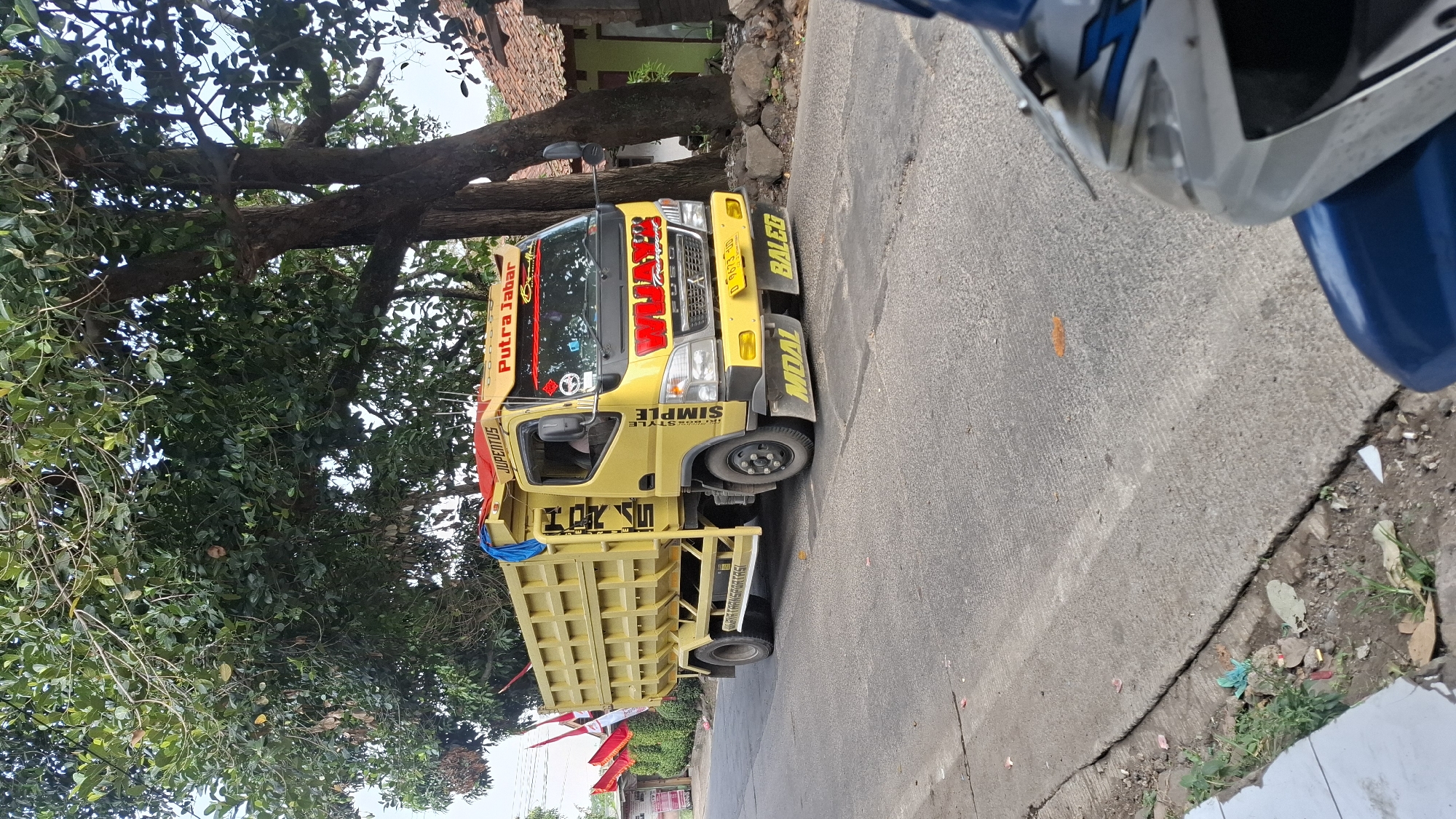 A yellow dump truck, identified as belonging to "PUTRA JABAR," is driving on a paved road in Cilaku, Indonesia. The truck has "JUPE NTUS" written above the windshield and "WUAT TRANSPORT" on the front grill. There are red and white flags attached to the truck's side. The truck is in motion, with its wheels turning. The environment includes a large tree with dense green foliage overhanging the road, and to the right, a house with a tiled roof. The road surface shows some cracks. In the foreground, a portion of a blue and silver motorcycle is visible, suggesting the image was taken from its perspective.